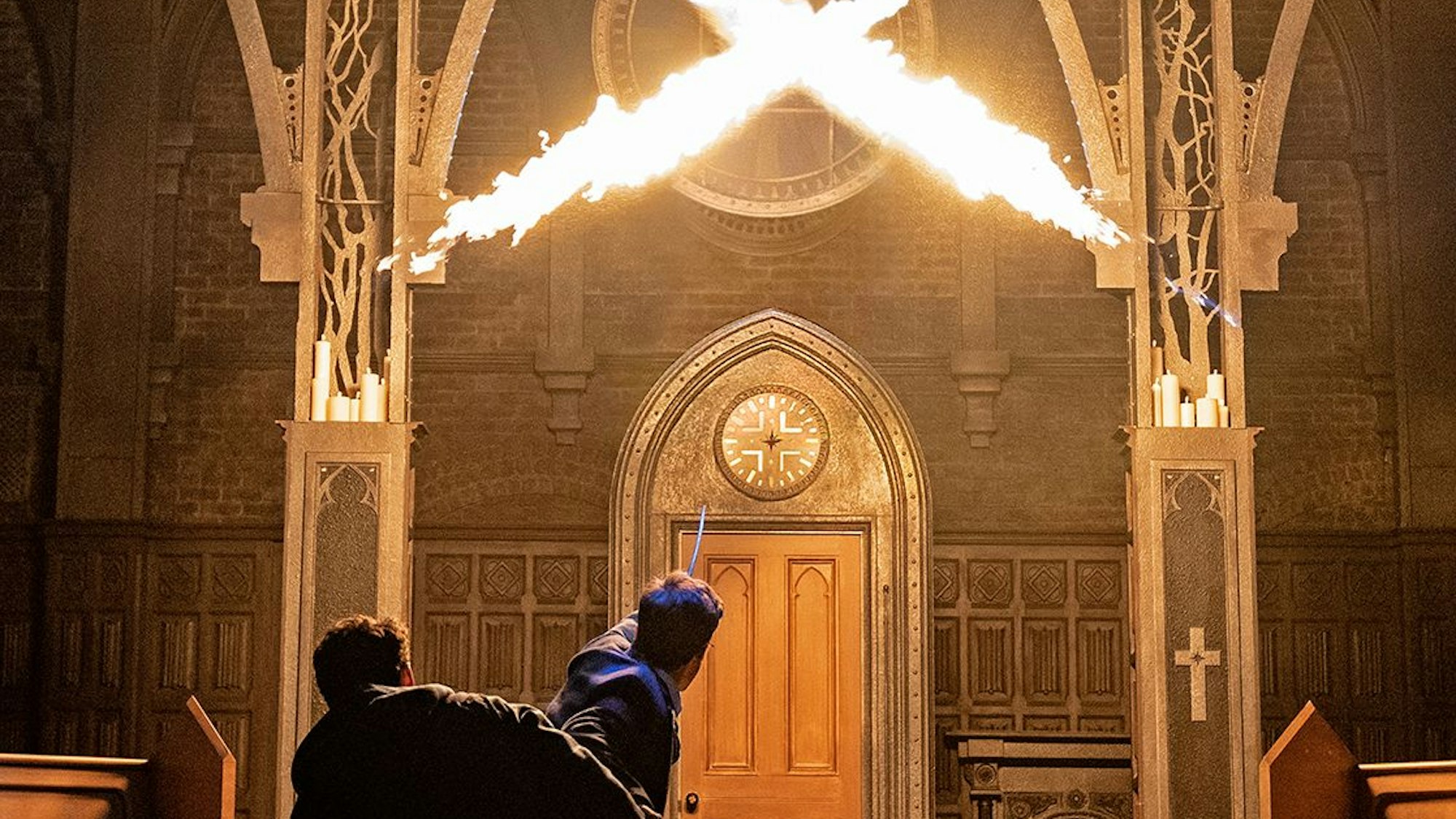 Die Charaktere Harry Potter, Albus Dumbledore Potter, Lucius Malfoy, Ginny Weasley, Hermione Granger und Ron Weasley halten ihre Zauberstäbe hoch, aus zweien kommen Feuerstöße, die sich in der Mitte der Bühne kreuzen.