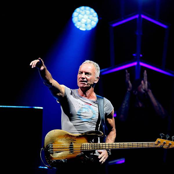 Sting steht im Rampenlicht, trägt ein graues T-Shirt und hat seine E-Gitarre umgehangen.