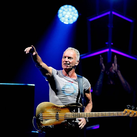 Sting steht im Rampenlicht, trägt ein graues T-Shirt und hat seine E-Gitarre umgehangen.