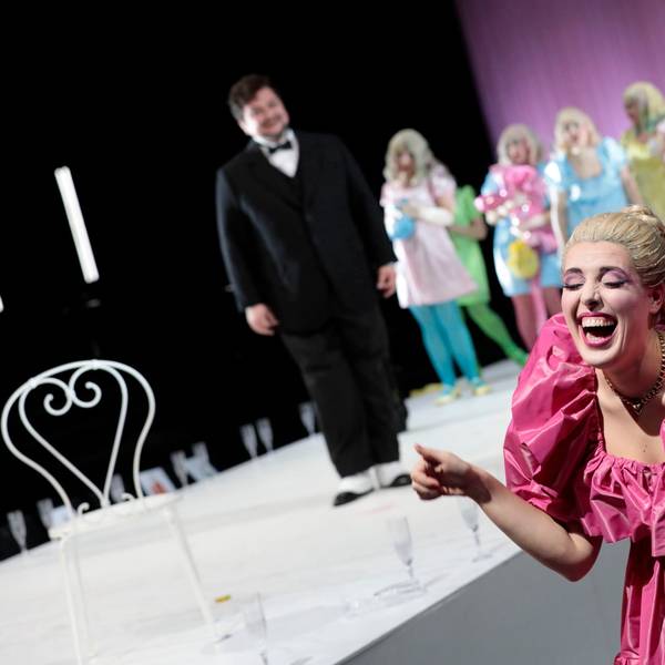 Eine Frau im rosa Rüschenkleid lacht schallen, im Hintergrund schaut die Titelfigur der Oper etwas ratlos drein.