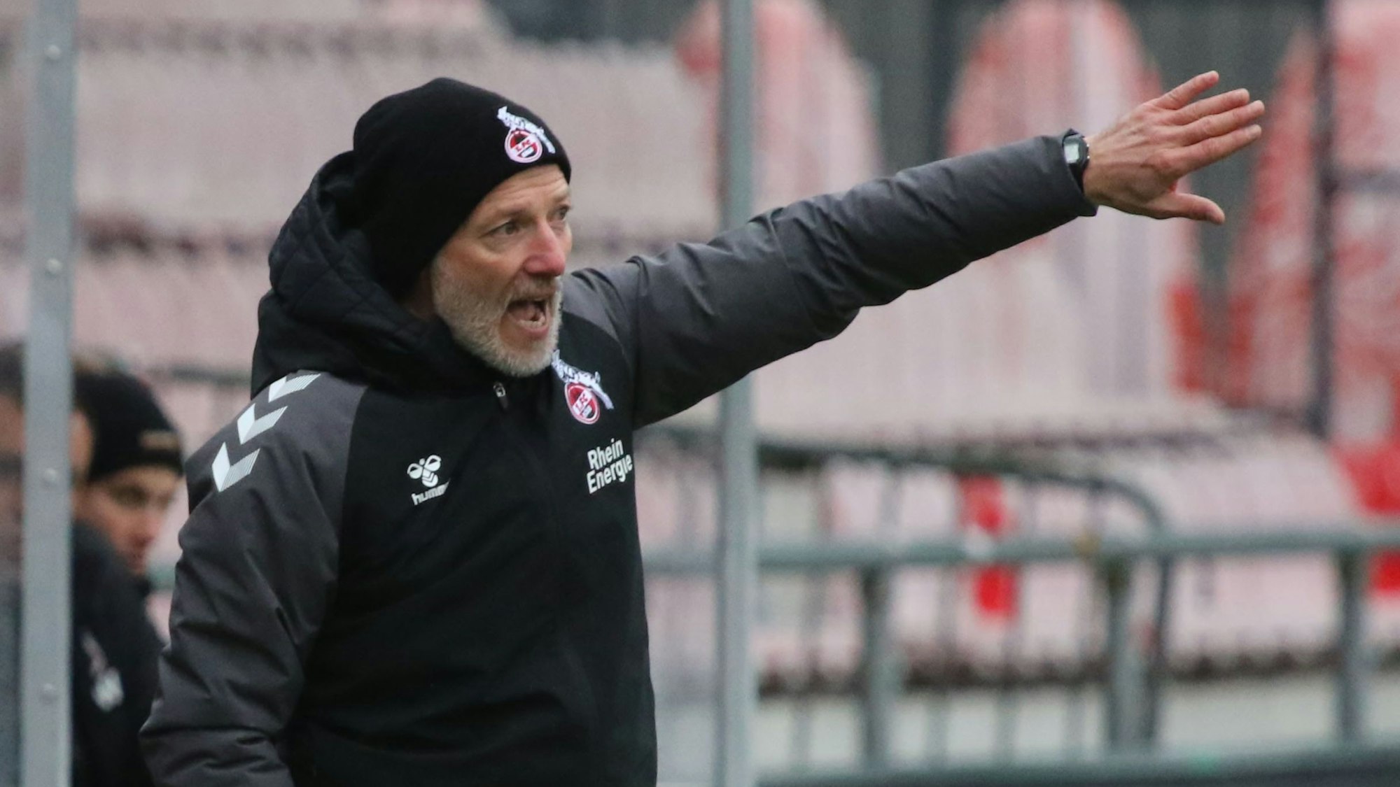 Ein Fußballtrainer steht am Spielfeldrand und gestikuliert mit der linken Hand.