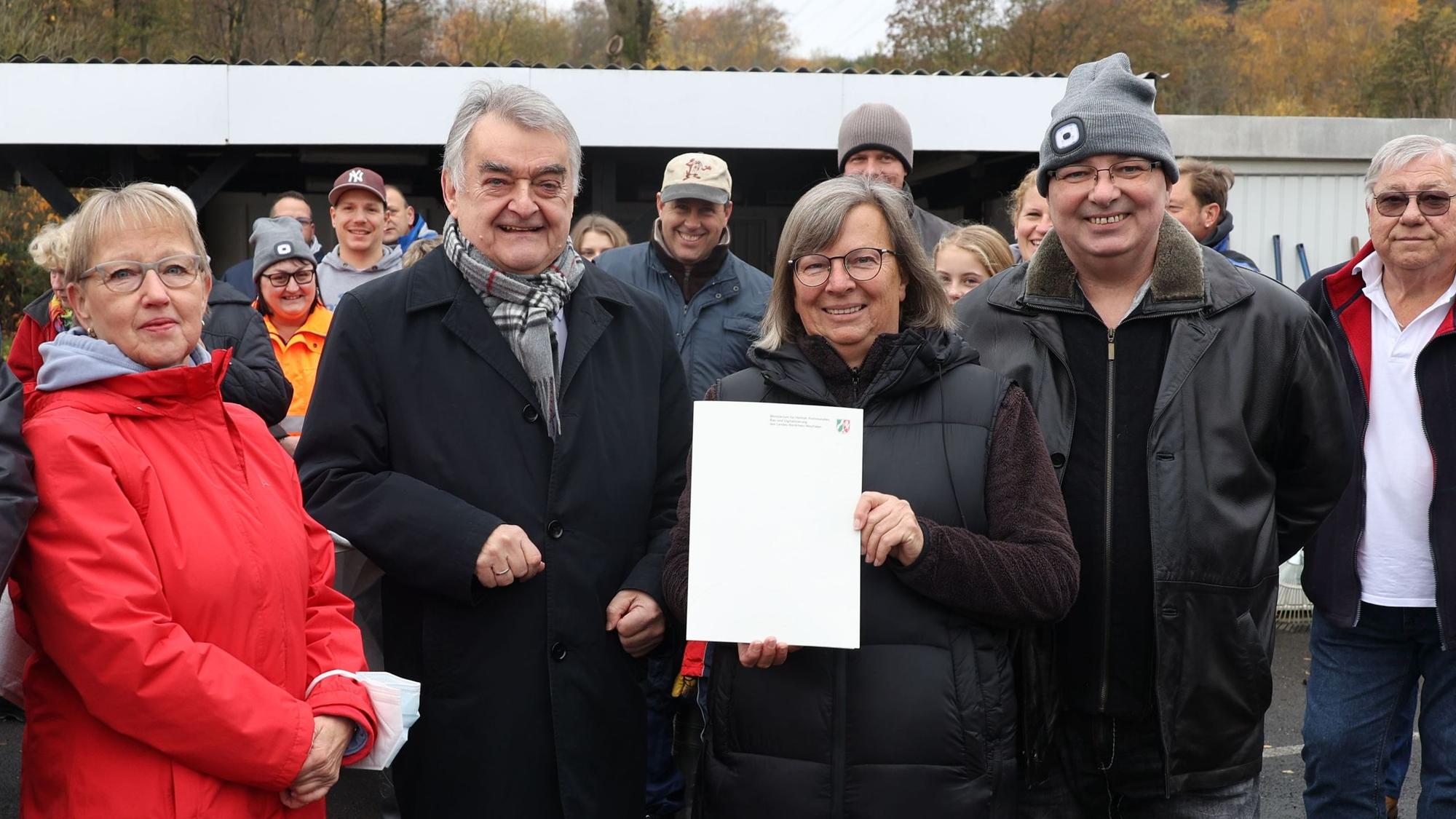 Innenminister Herbert Reul steht mit Kassiererin Dagmar Sapina umringt von Clubmitgliedern auf dem Campingplatz und übergibt den schriftlichen Bescheid über eine halbe Million Euro aus dem Fluthilfe-Fonds.