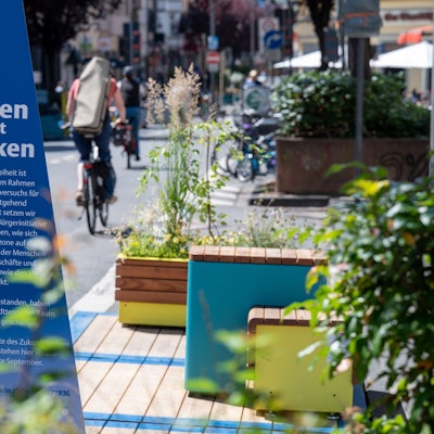Zu sehen sind Sitzbänke auf der Deutzer Freiheit im Jahr 2022, die die Stadt Köln unter dem Motto „Sitzen statt Parken“ auf Parkplätzen aufgestellt hatte, sowie ein großes blaues Schild mit der Aufschrift „Sitzen statt Parken“.