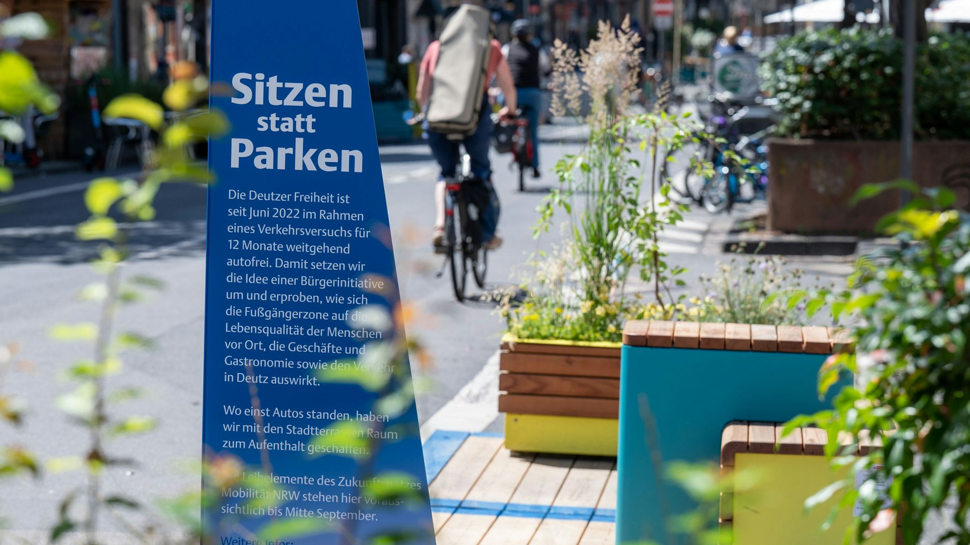 Zu sehen sind Sitzbänke auf der Deutzer Freiheit im Jahr 2022, die die Stadt Köln unter dem Motto „Sitzen statt Parken“ auf Parkplätzen aufgestellt hatte, sowie ein großes blaues Schild mit der Aufschrift „Sitzen statt Parken“.