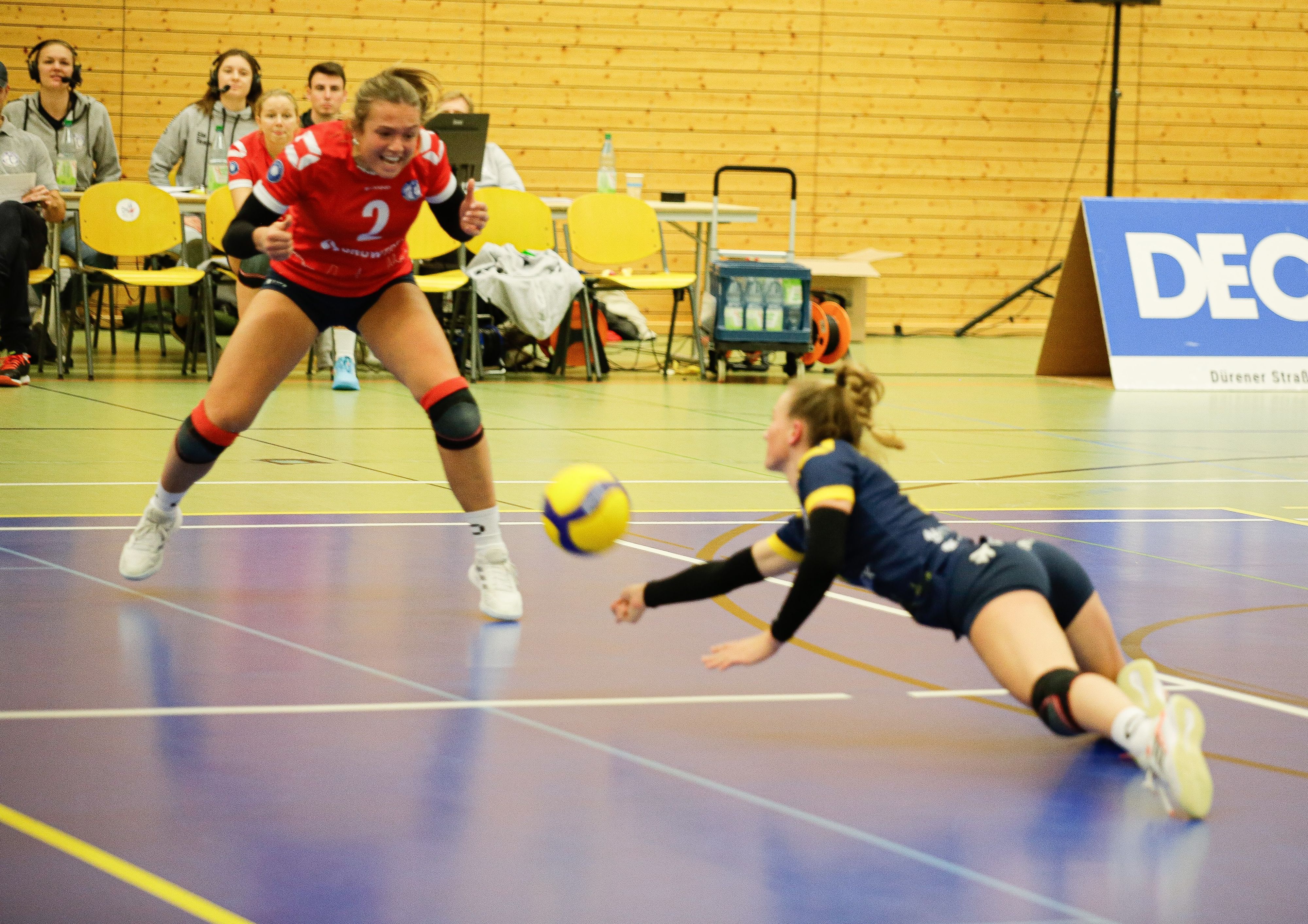 Eine Volleyballspielerin wirft sich im Kampf um den Ball auf den Boden. Eine Mitspielerin steht vor ihr und feuert sie an.