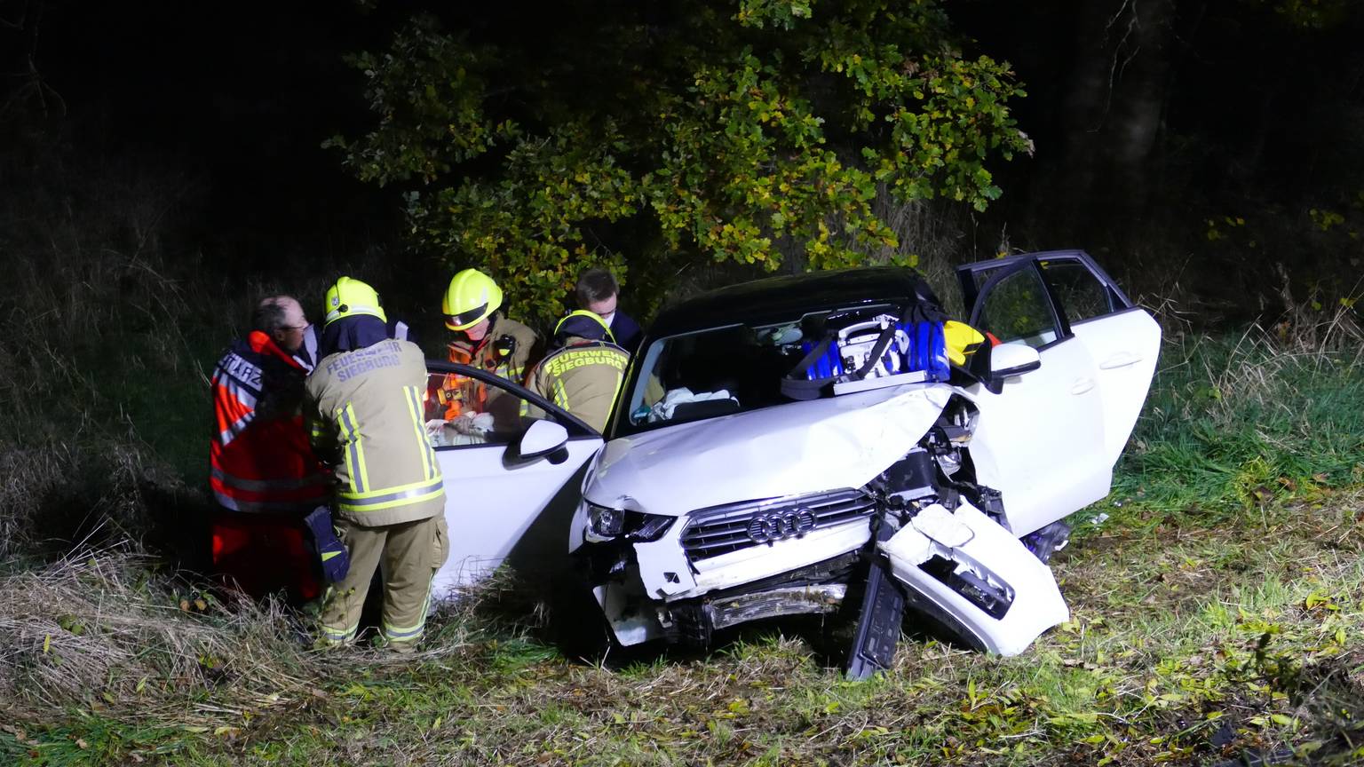 Ein weißes Auto steht im Graben, die Türen stehen offen, das Heck ist demoliert. Daneben stehen Feuerwehrleute und ein Notarzt.