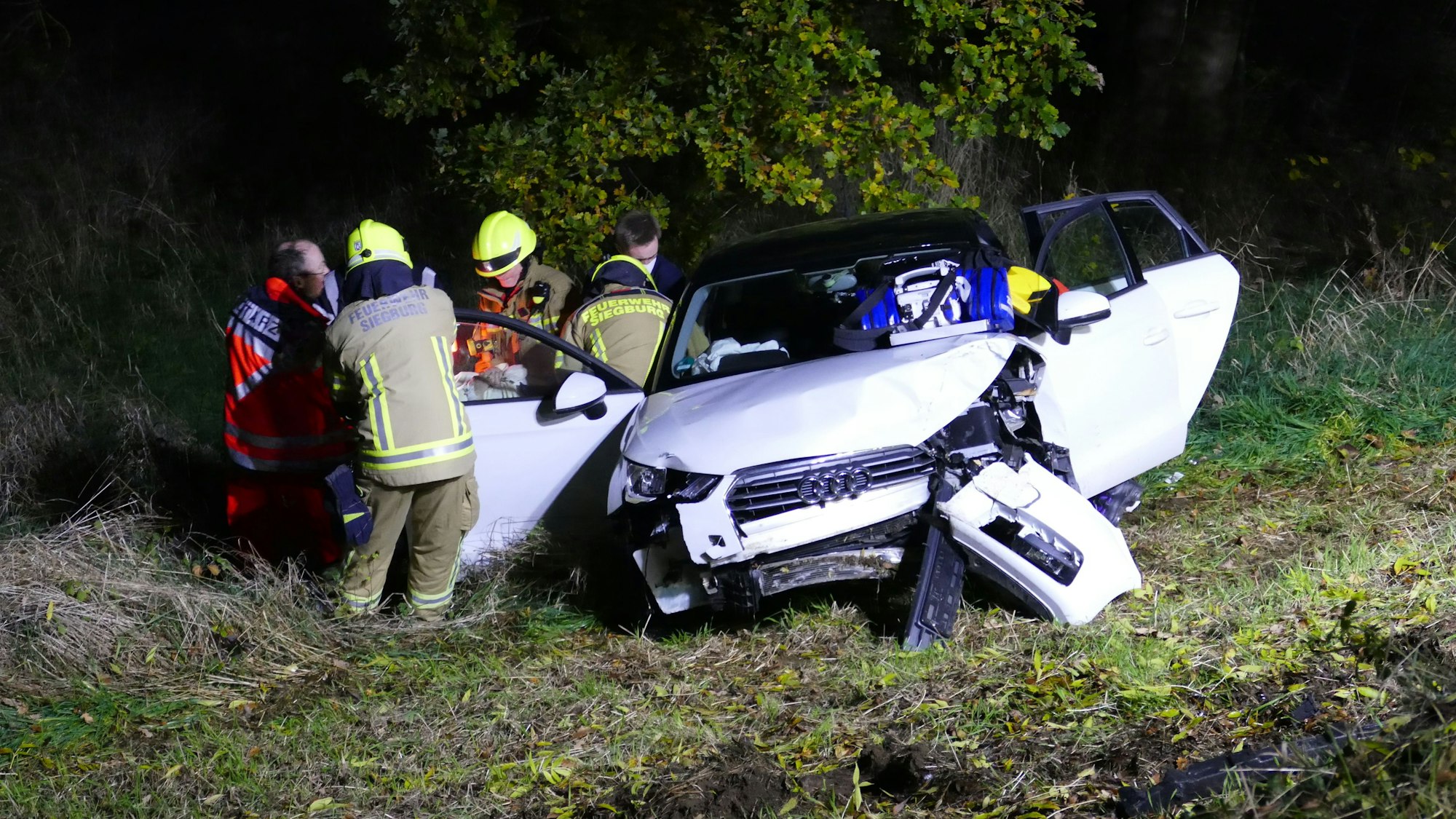 Ein weißes Auto steht im Graben, die Türen stehen offen, das Heck ist demoliert. Daneben stehen Feuerwehrleute und ein Notarzt.