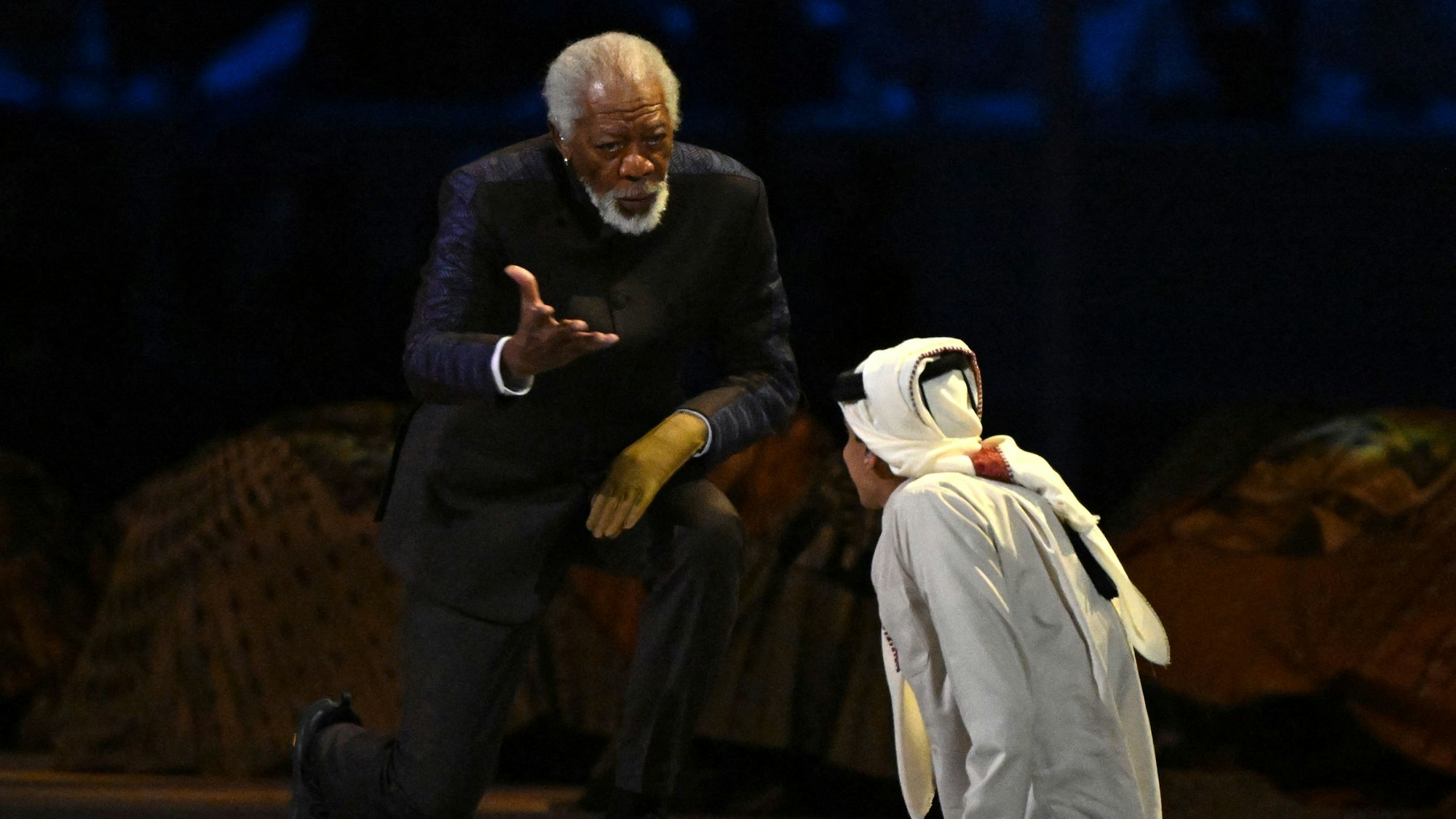 Morgan Freeman (l.) und Paralympics-Sportler Ghanim Al-Muftah bei ihrem Auftritt an der WM-Eröffnungsfeier. Freeman spricht und streckt seine Hand zum Sportler aus. Mit der anderen Hand stützt er sich auf dem Knie ab.
