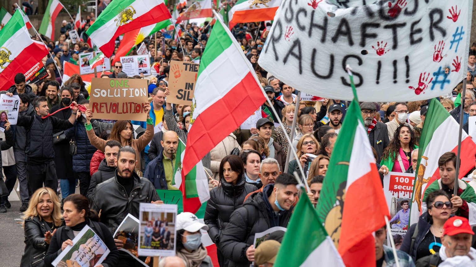 Bei einer Demonstration laufen zahlreiche Menschen mit dem Demozug mit und halten Iran-Flaggen in die Luft.