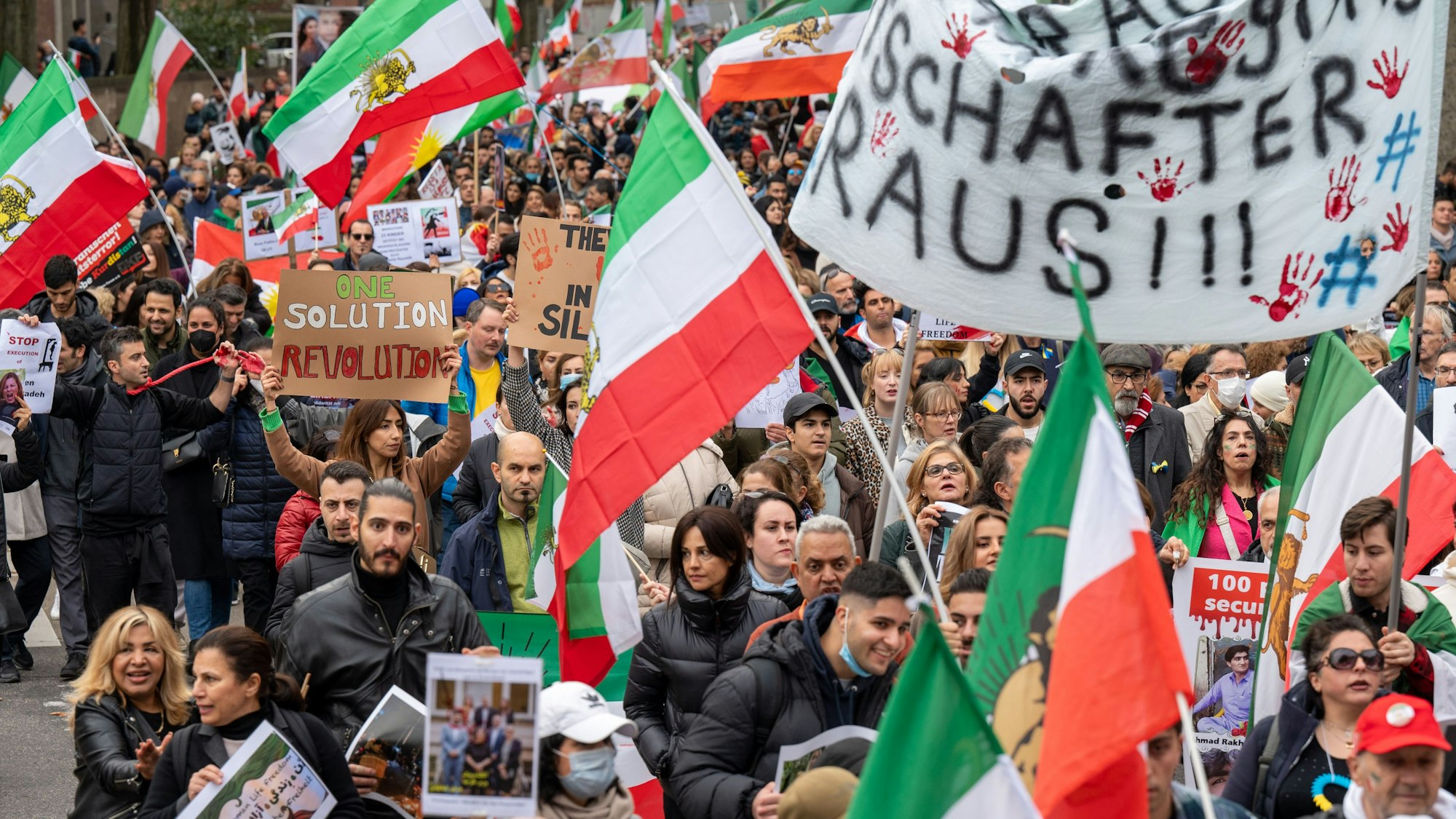 Bei einer Demonstration laufen zahlreiche Menschen mit dem Demozug mit und halten Iran-Flaggen in die Luft.