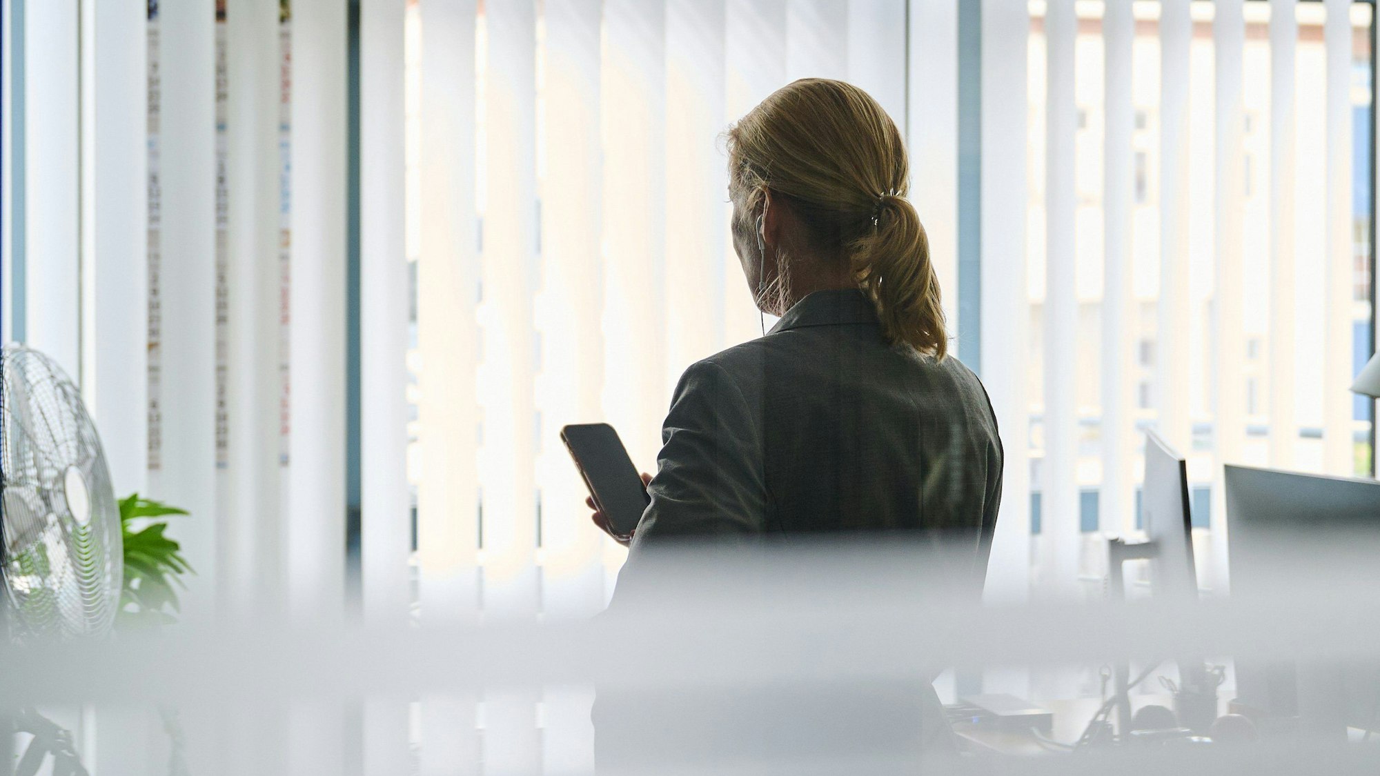 Eine Frau steht in einem Bürozimmer am Fenster und telefoniert über ein Headset (gestellte Szene).