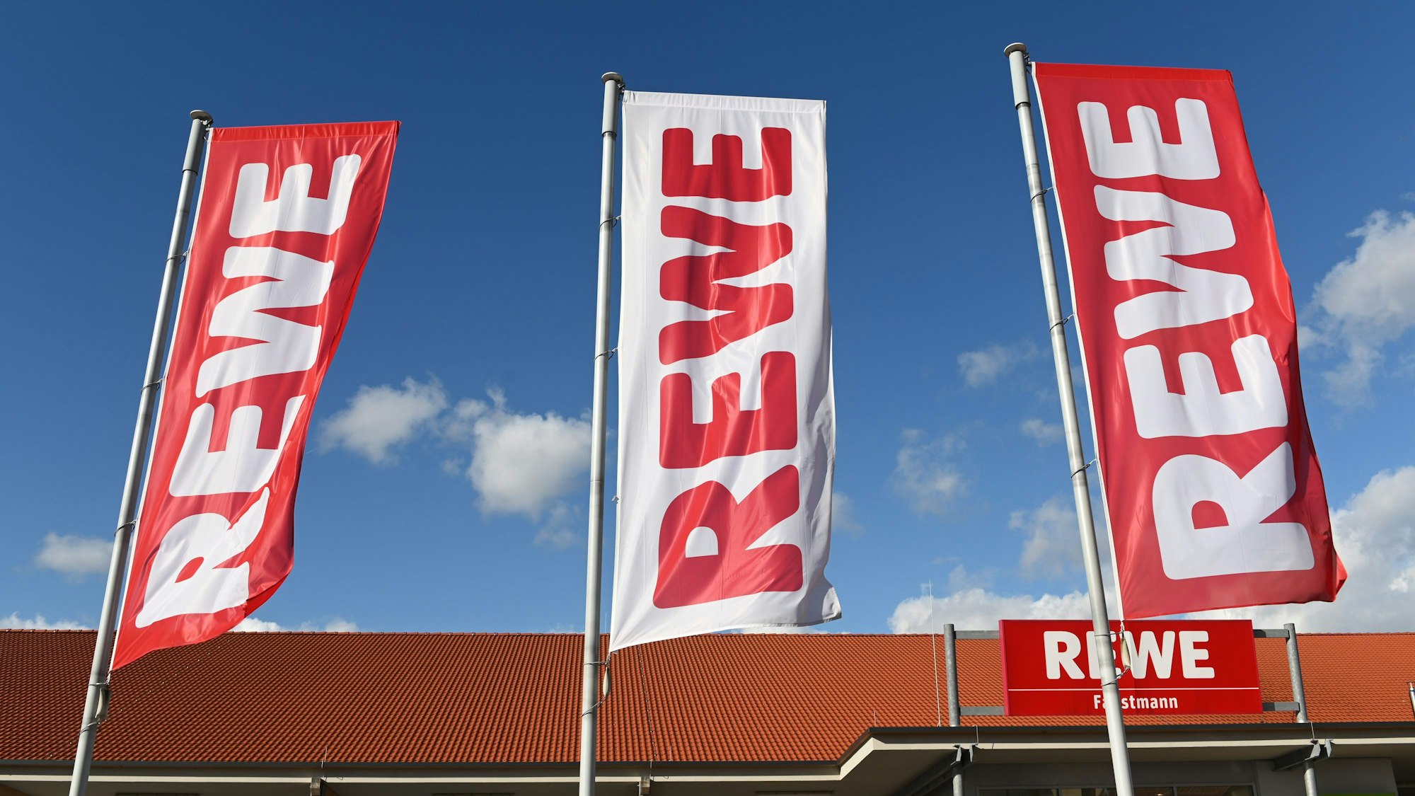 Es sind drei Rewe-Flaggen vor dem Supermarkt zu sehen.