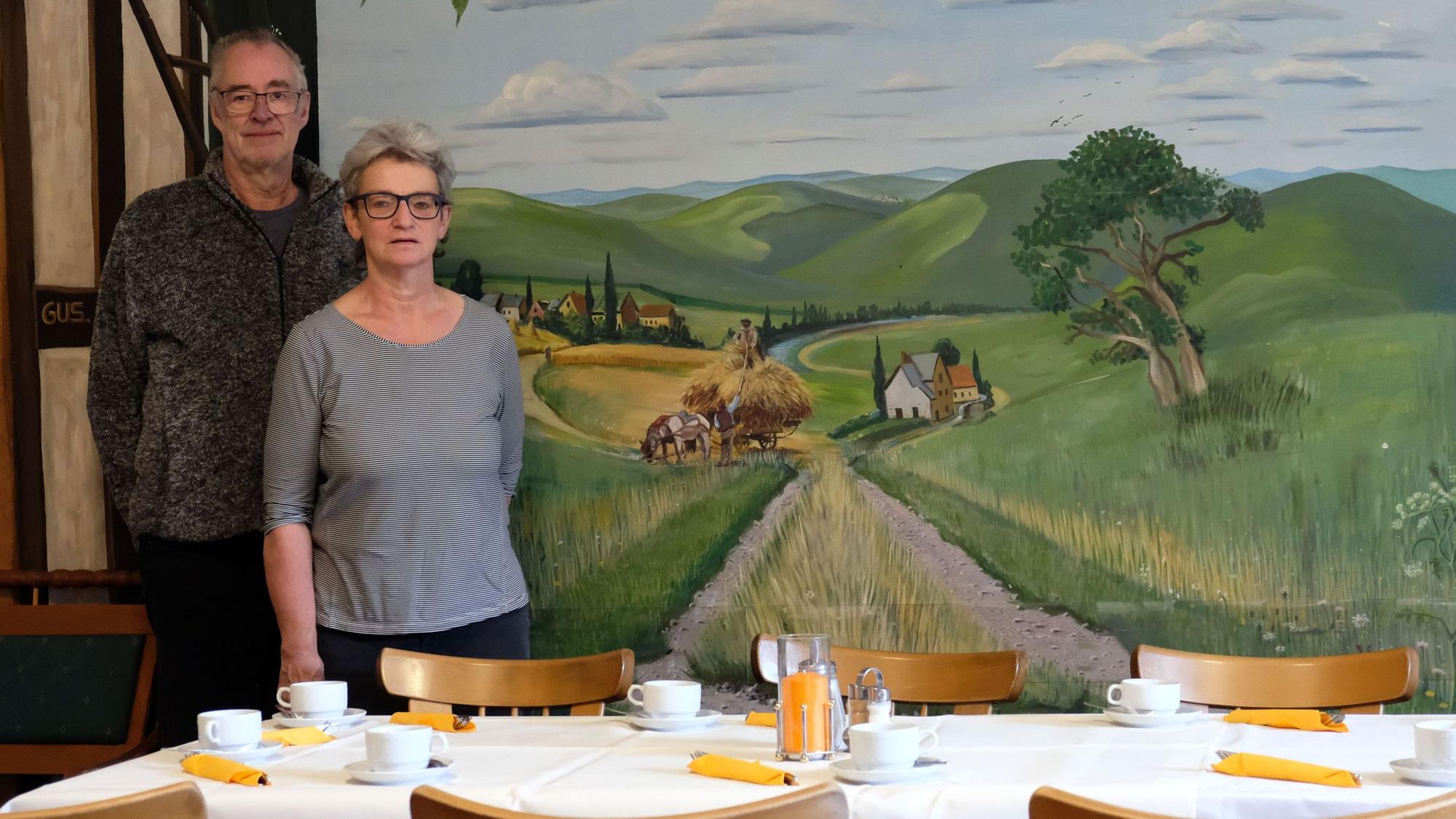 Annegret und Volker Prause, die seit 2004 den "Brunnenhof" in Mechernich-Holzheim betrieben haben, stehen vor einem Tisch, hinter ihnen ist ein Feldweg auf eine Wand gemalt.