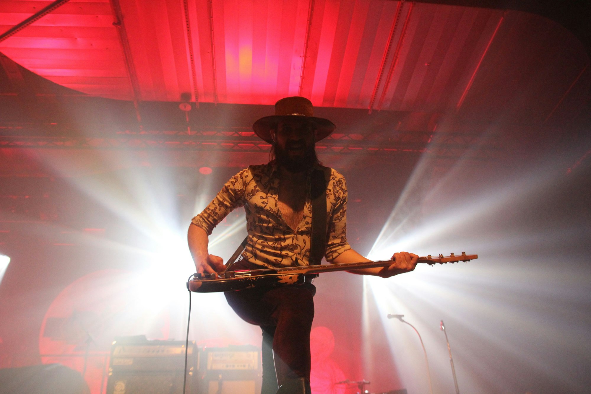 Der Bassist Simon Bouteloup hält sein Instrument quer vor sich. Er wird von weißen Lichtstrahlen flankiert, während sein Gesicht im Schatten seiner Hutkrempe verborgen ist.