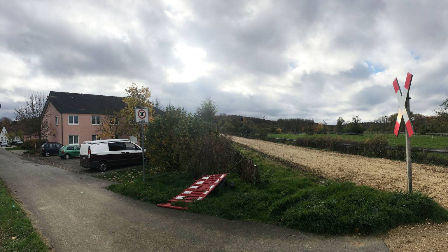 Auf der Bahnstrecke liegen keine Gleise. Neben der Strecke liegt ein Haus. Davor stehen Autos.