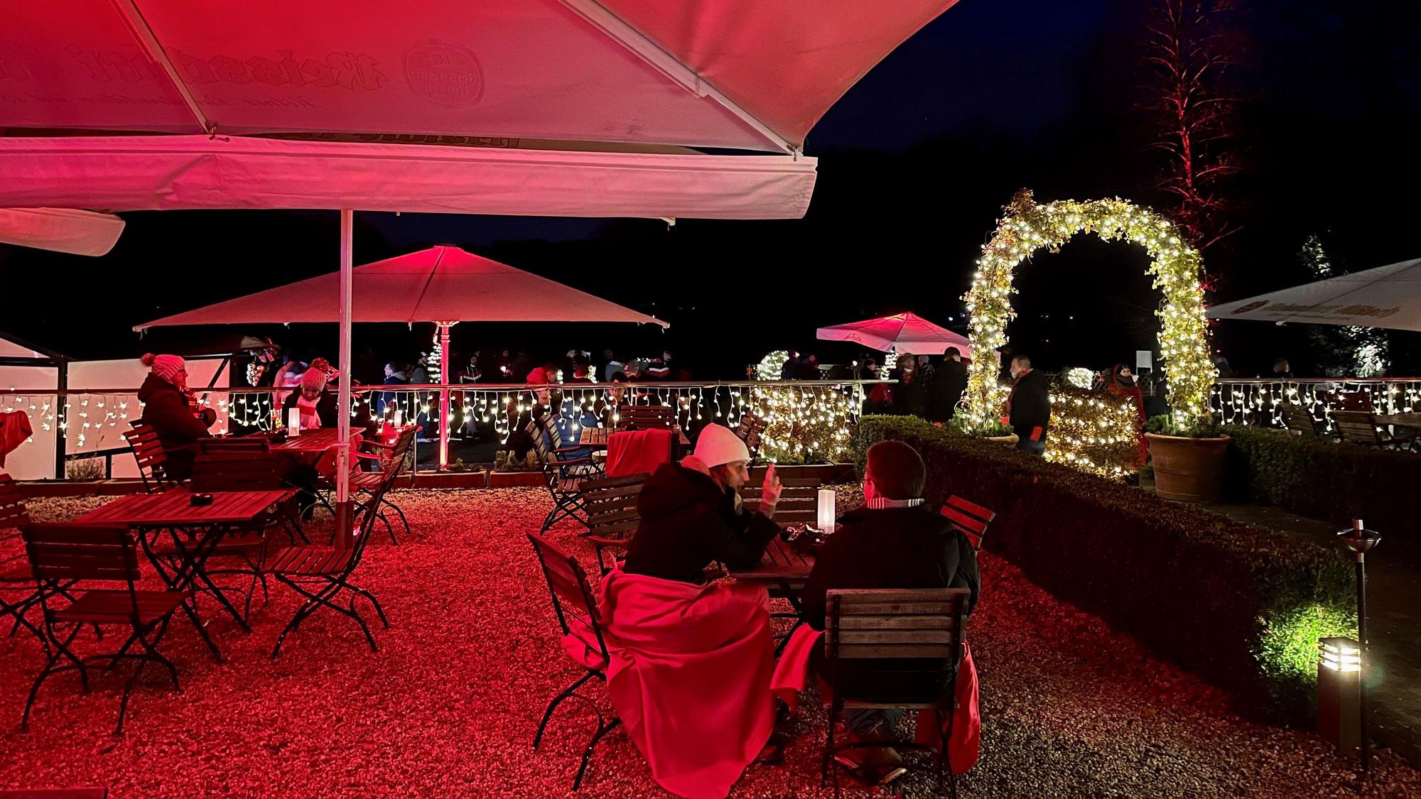 auf einer Terrasse sitzen unter großen Schirmen Menschen mit Decken an Holztischen, im Hintergrund weihnachtliche Beleuchtung in der Dunkelheit