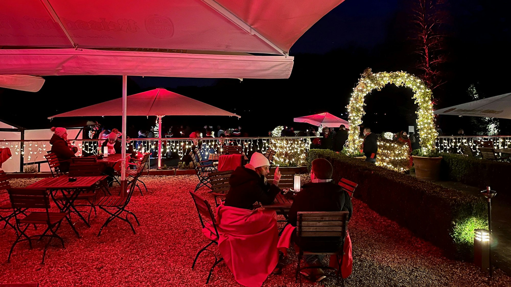 auf einer Terrasse sitzen unter großen Schirmen Menschen mit Decken an Holztischen, im Hintergrund weihnachtliche Beleuchtung in der Dunkelheit