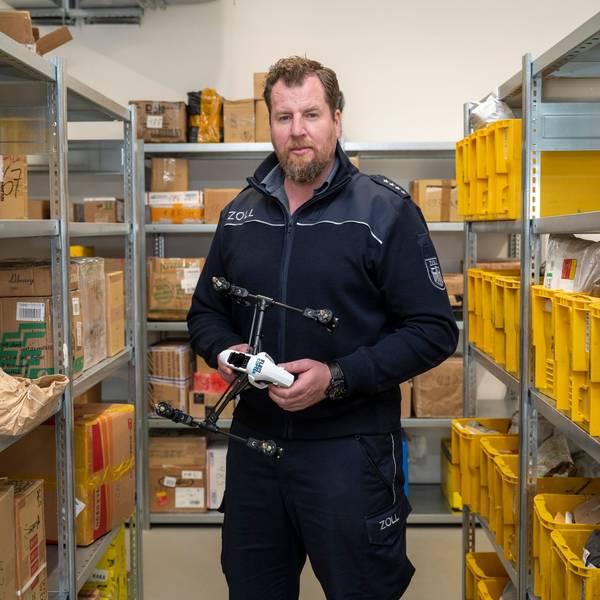 Zoll-Sprecher Jens Ahland steht zwischen Regalen mit Paketen, die der Zoll aus dem Verkehr gefischt hat, und hält eine Drohne in der Hand, die in Deutschland nicht zugelassen ist.
