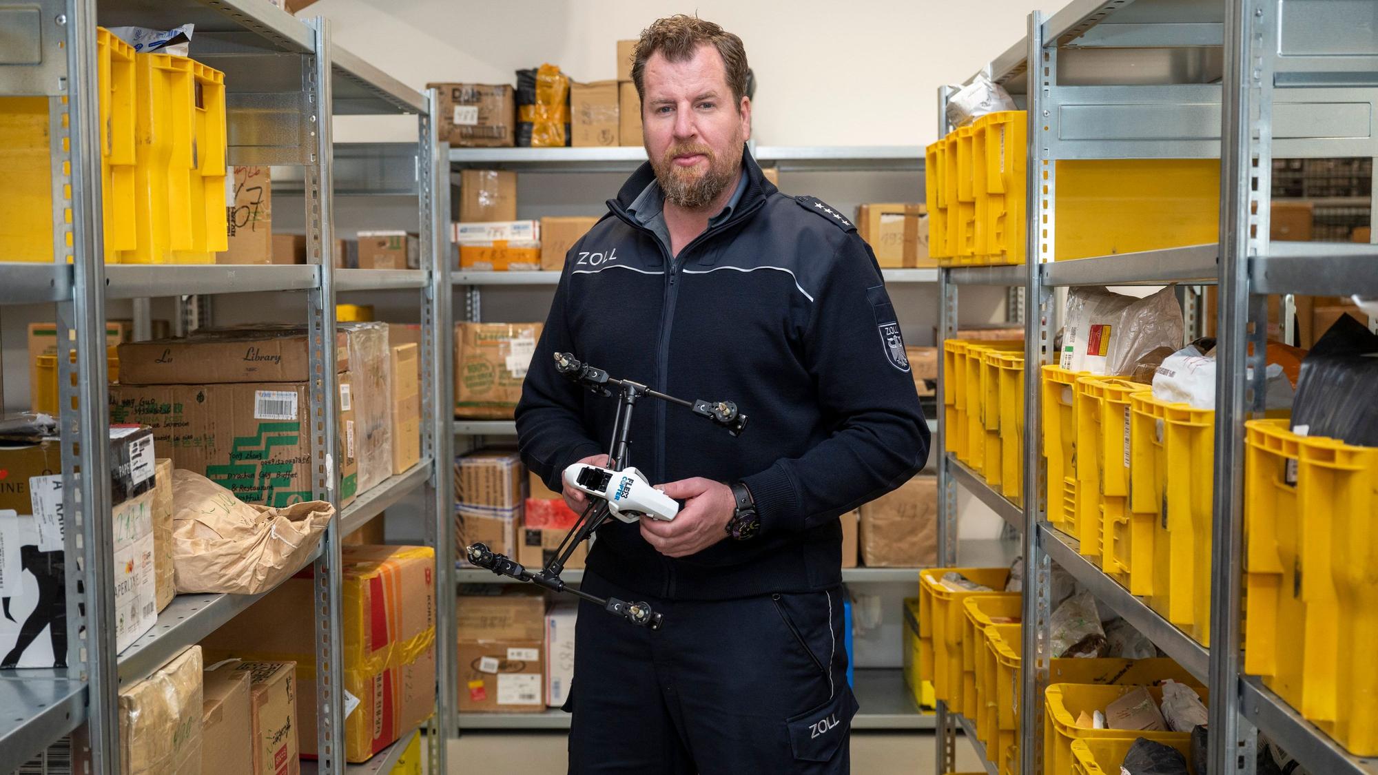 Zoll-Sprecher Jens Ahland steht zwischen Regalen mit Paketen, die der Zoll aus dem Verkehr gefischt hat, und hält eine Drohne in der Hand, die in Deutschland nicht zugelassen ist.