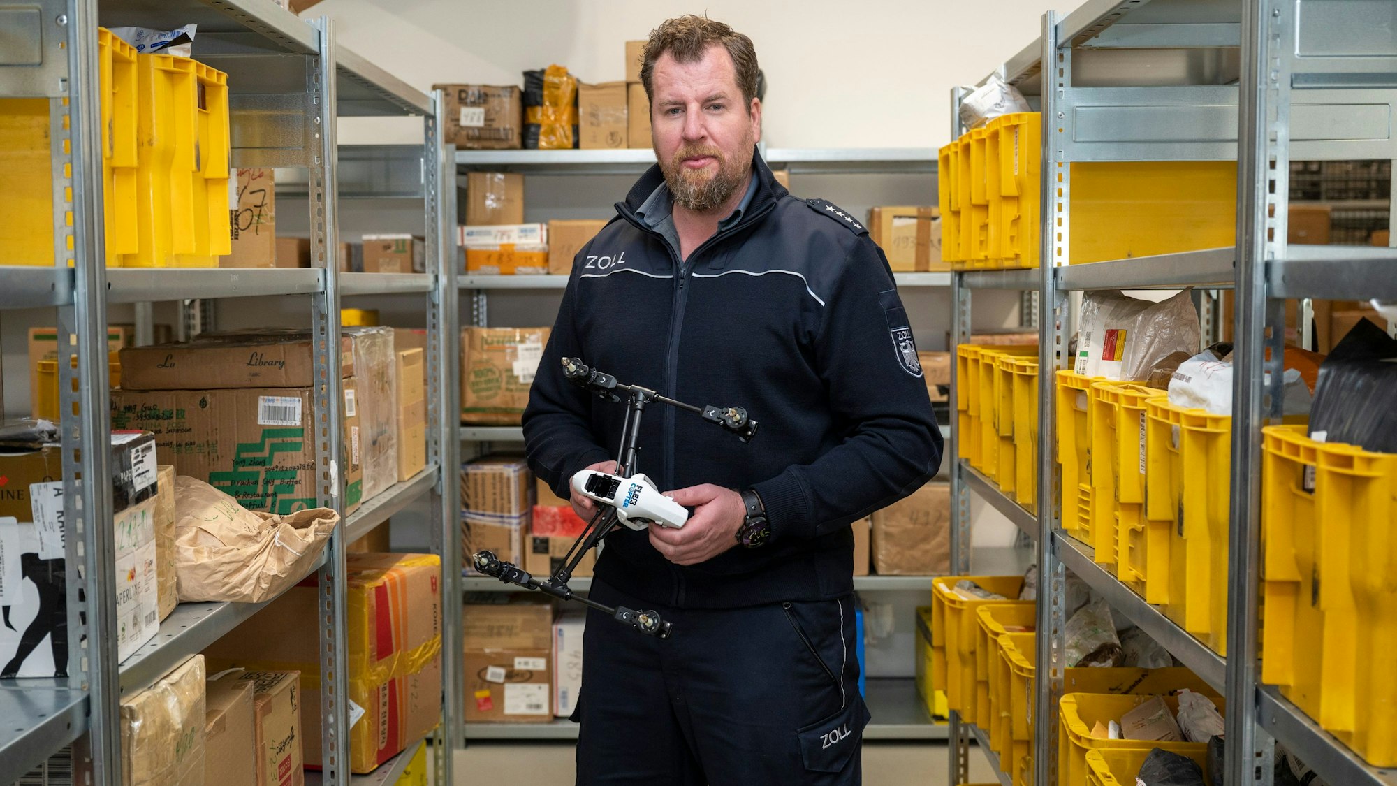 Zoll-Sprecher Jens Ahland steht zwischen Regalen mit Paketen, die der Zoll aus dem Verkehr gefischt hat, und hält eine Drohne in der Hand, die in Deutschland nicht zugelassen ist.