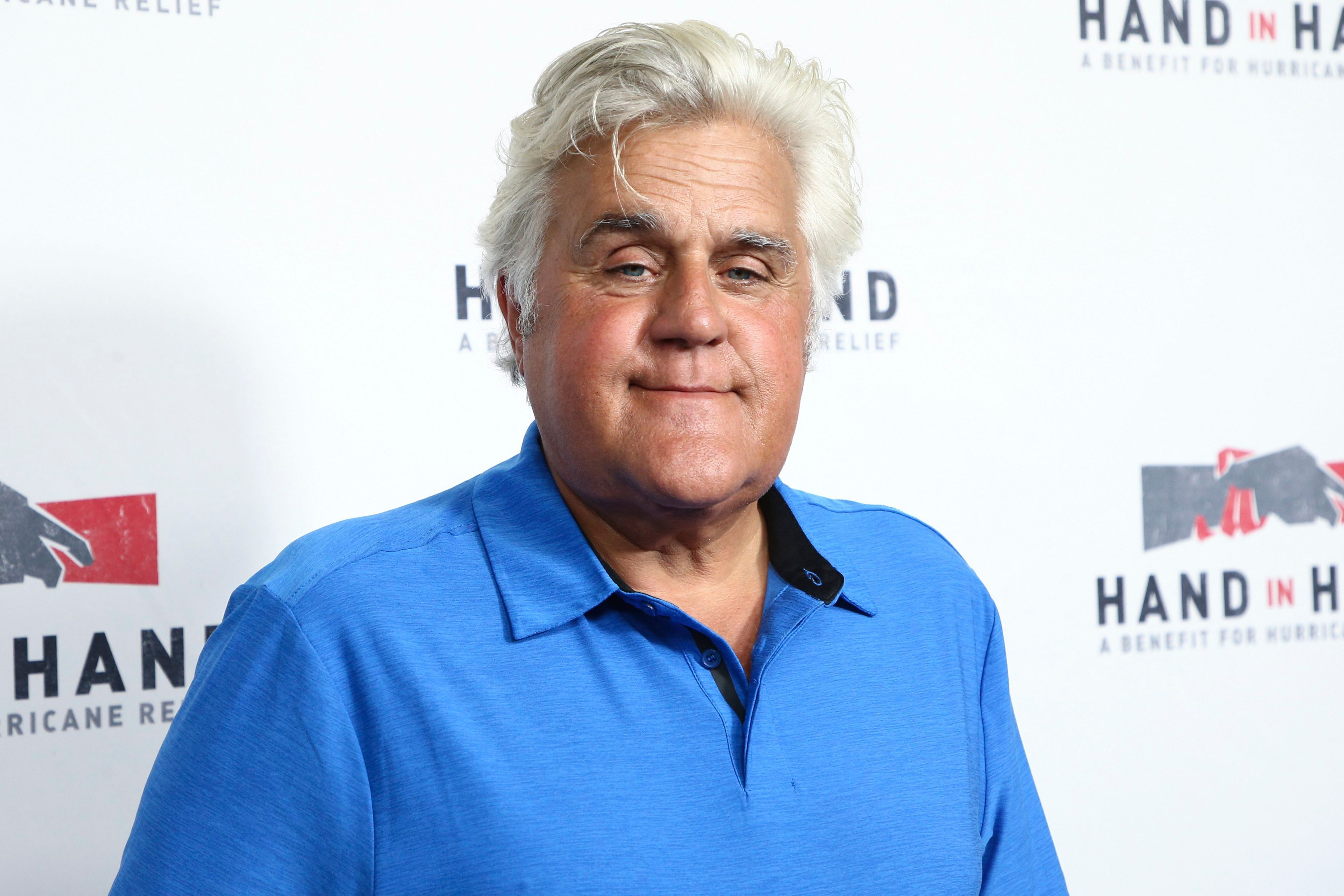 Der US-Moderator Jay Leno steht in einem blauen Polohemd vor einer weißen Wand.