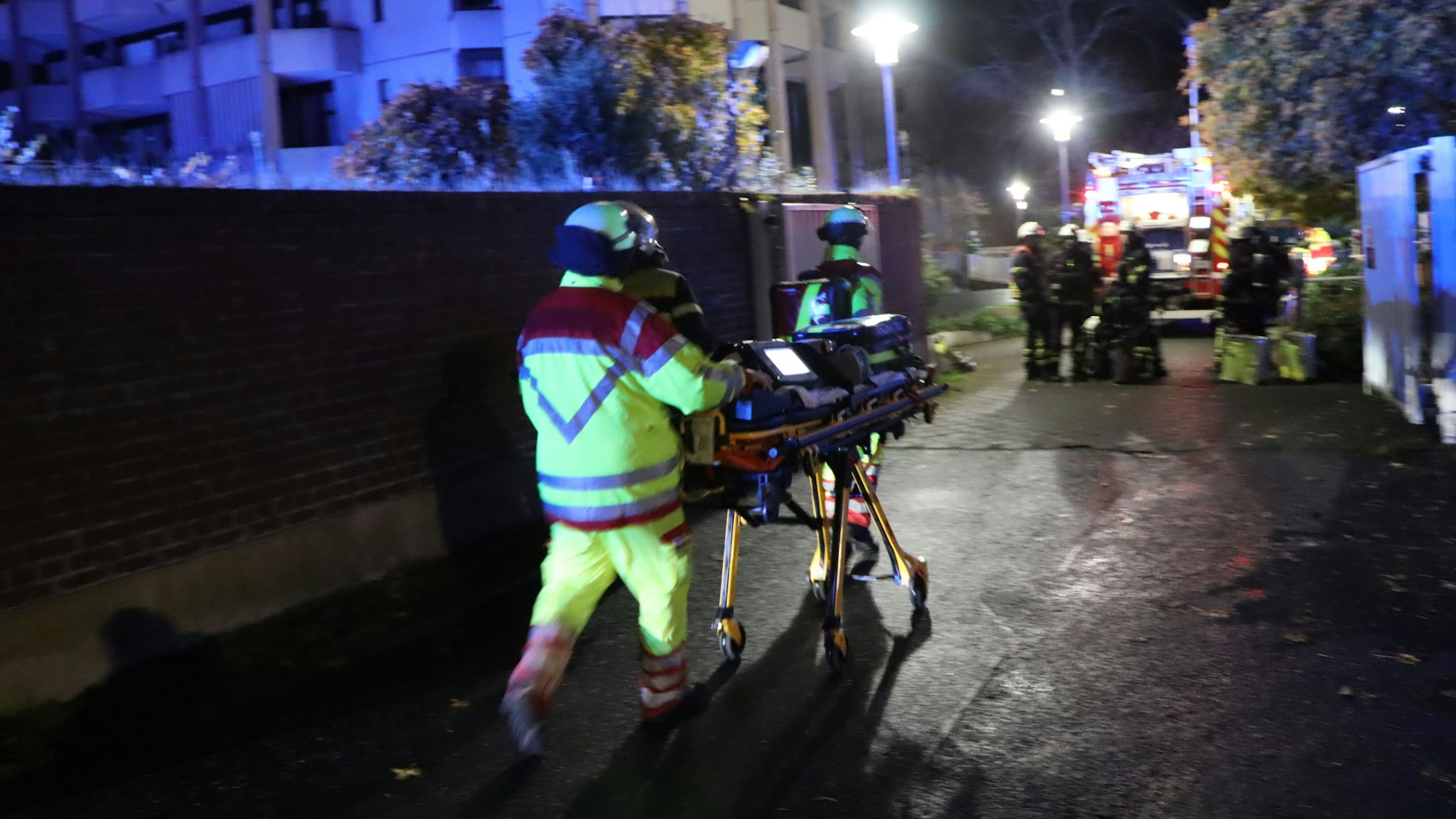 Einsatzkräfte der Feuerwehr schieben eine Trage über die Straße. Es ist Nacht, die Szene ist von Blaulicht erleuchtet.