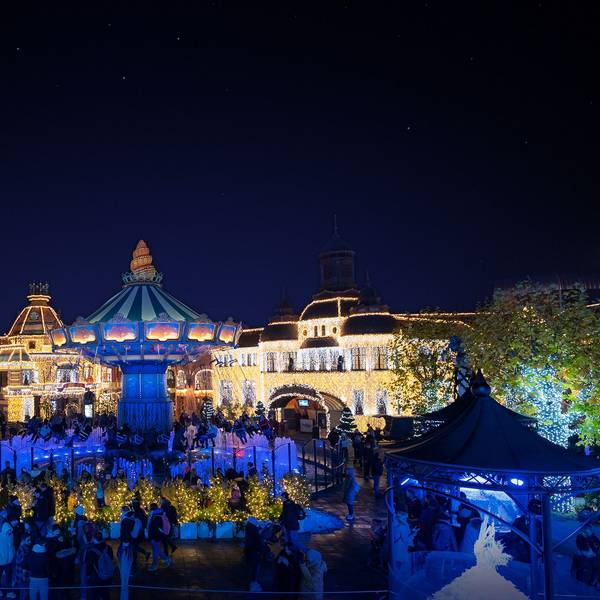 Die Gebäude im Hintergrund sind mit vielen Lichtern beleuchtet, das Kettenkarussell leuchtet blau, viele Besucher stehen davor.