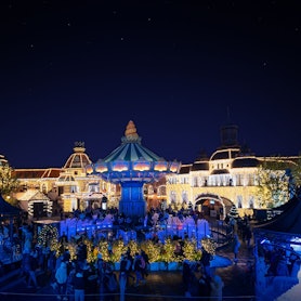 Die Gebäude im Hintergrund sind mit vielen Lichtern beleuchtet, das Kettenkarussell leuchtet blau, viele Besucher stehen davor.