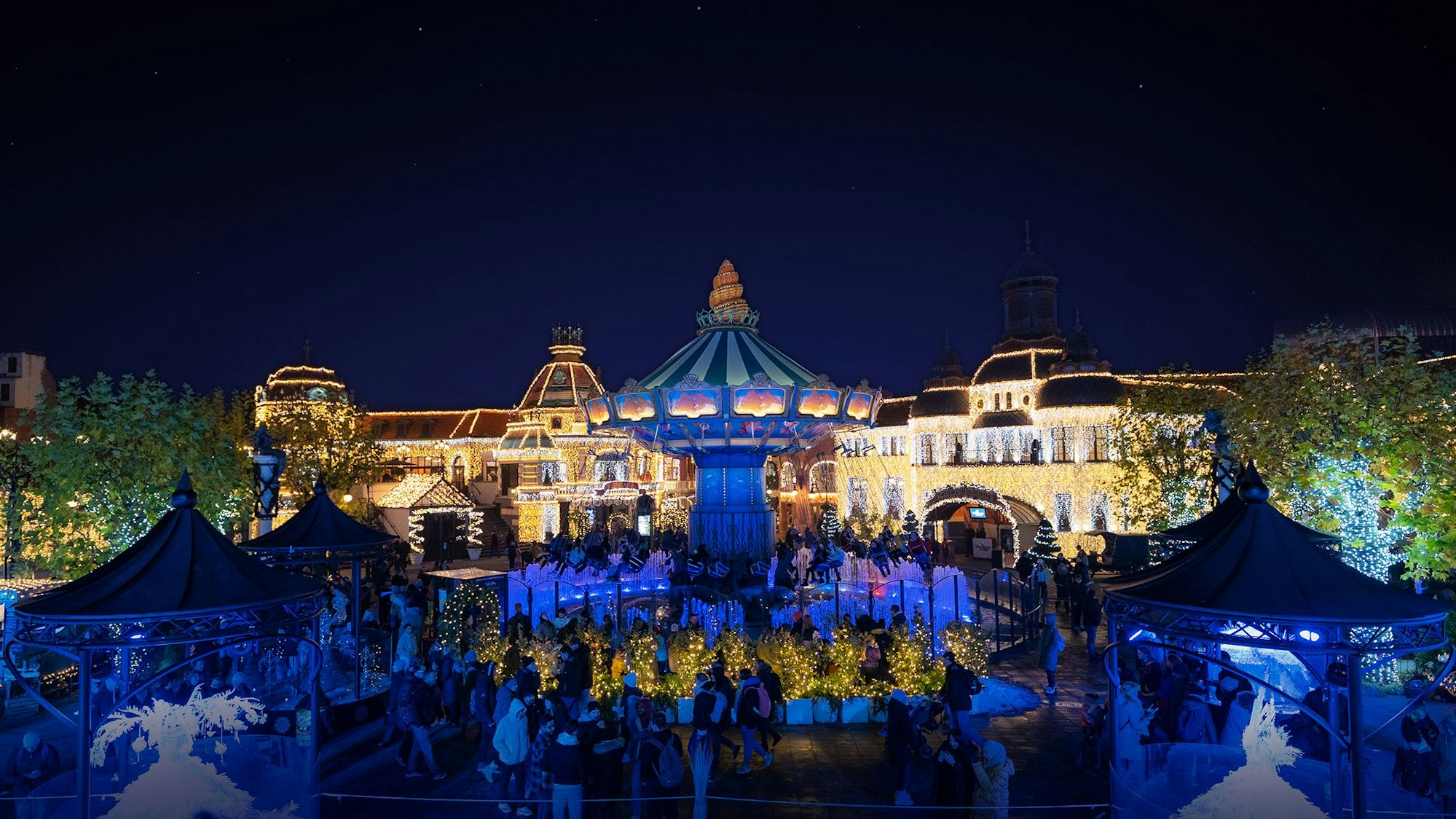 Die Gebäude im Hintergrund sind mit vielen Lichtern beleuchtet, das Kettenkarussell leuchtet blau, viele Besucher stehen davor.