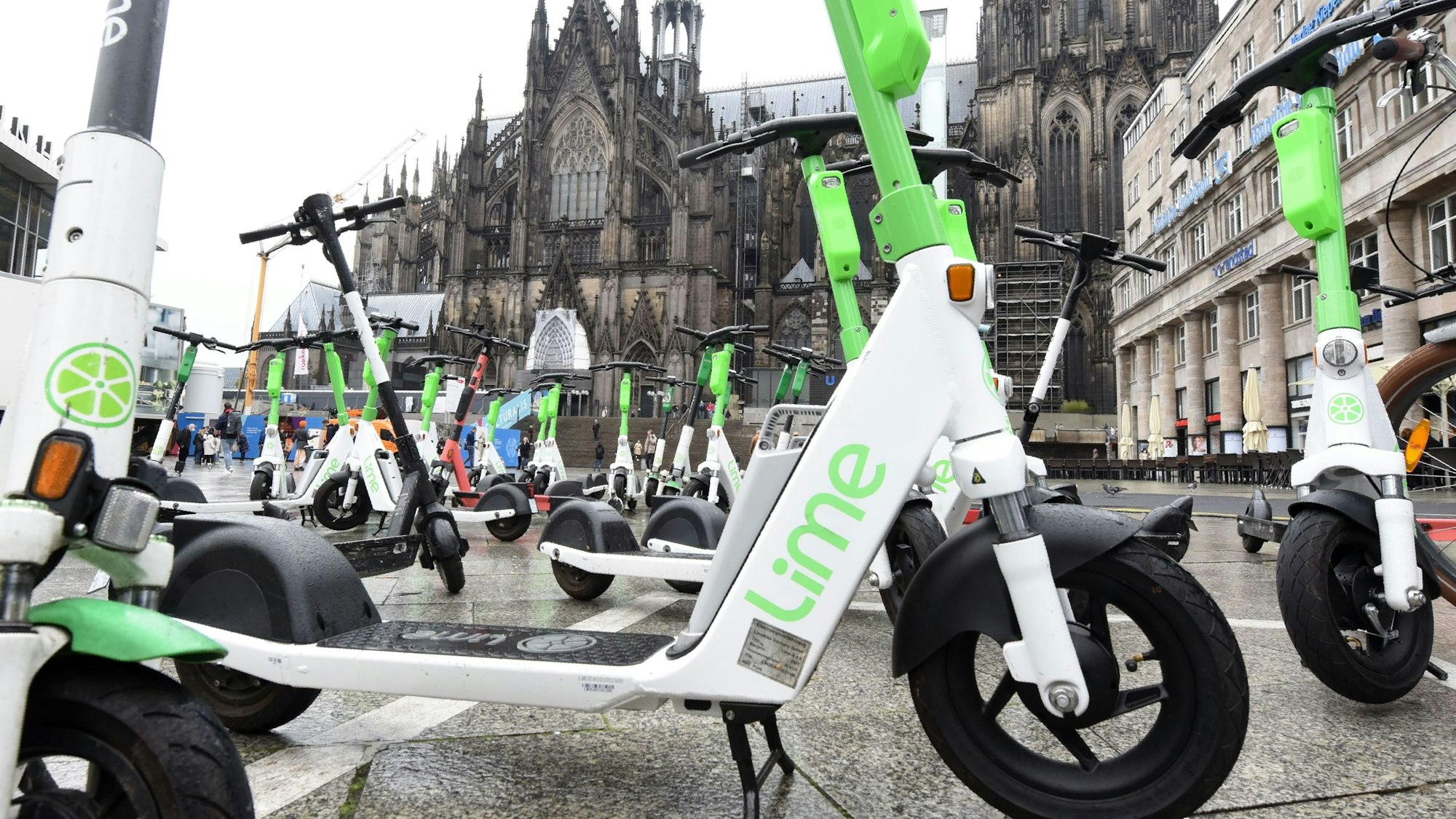 Elektroroller der Firma Lime stehen auf dem Bahnhofsvorplatz mit dem Dom im Hintergrund.
