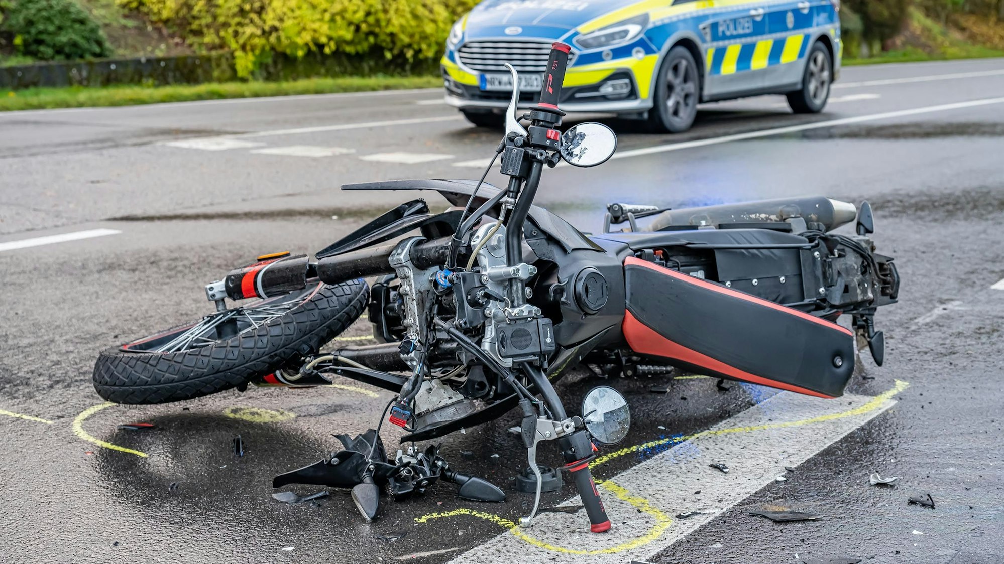 Ein Motorrad liegt beschädigt auf dem Boden. Die Umrisse sind mit gelber Farbe auf den Boden gemalt. Im Hintergrund steht ein Polizeiauto.