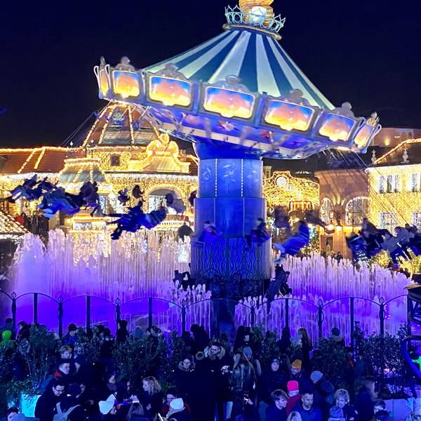 Zu sehen ist ein in bunten Farben beleuchtetes Kettenkarussell im Phantasialand sowie mit Lichter geschmückte Häuser im Hintergrund.