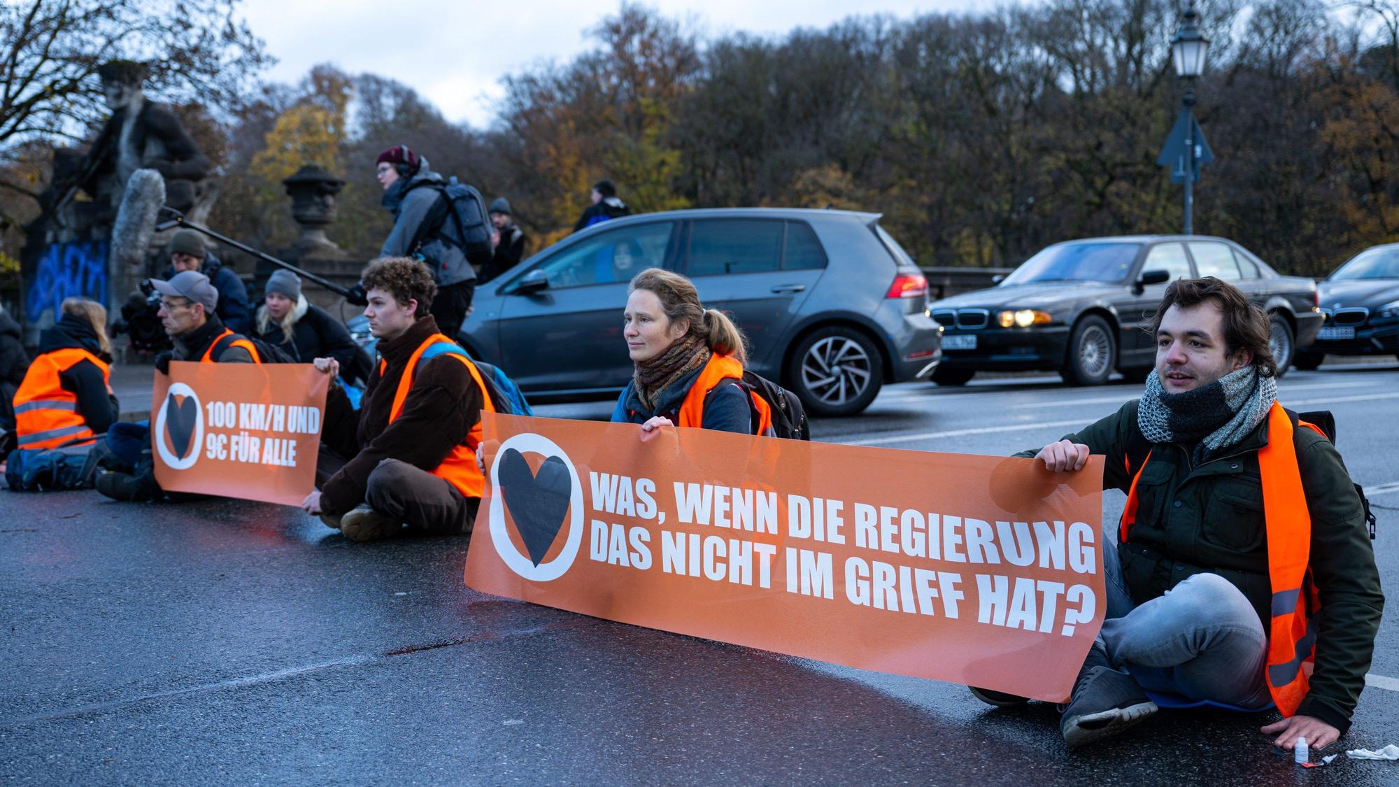 Klimaaktivisten der Umweltschutzbewegung „Letzte Generation“ sitzen auf der Prinzregentenstraße in München und blockieren den Verkehr. Sie halten Banner mit dn Aufschriften „Was, wenn die Regierung das nicht im Griff hat“ und „100 km/h und 9 € für alle“