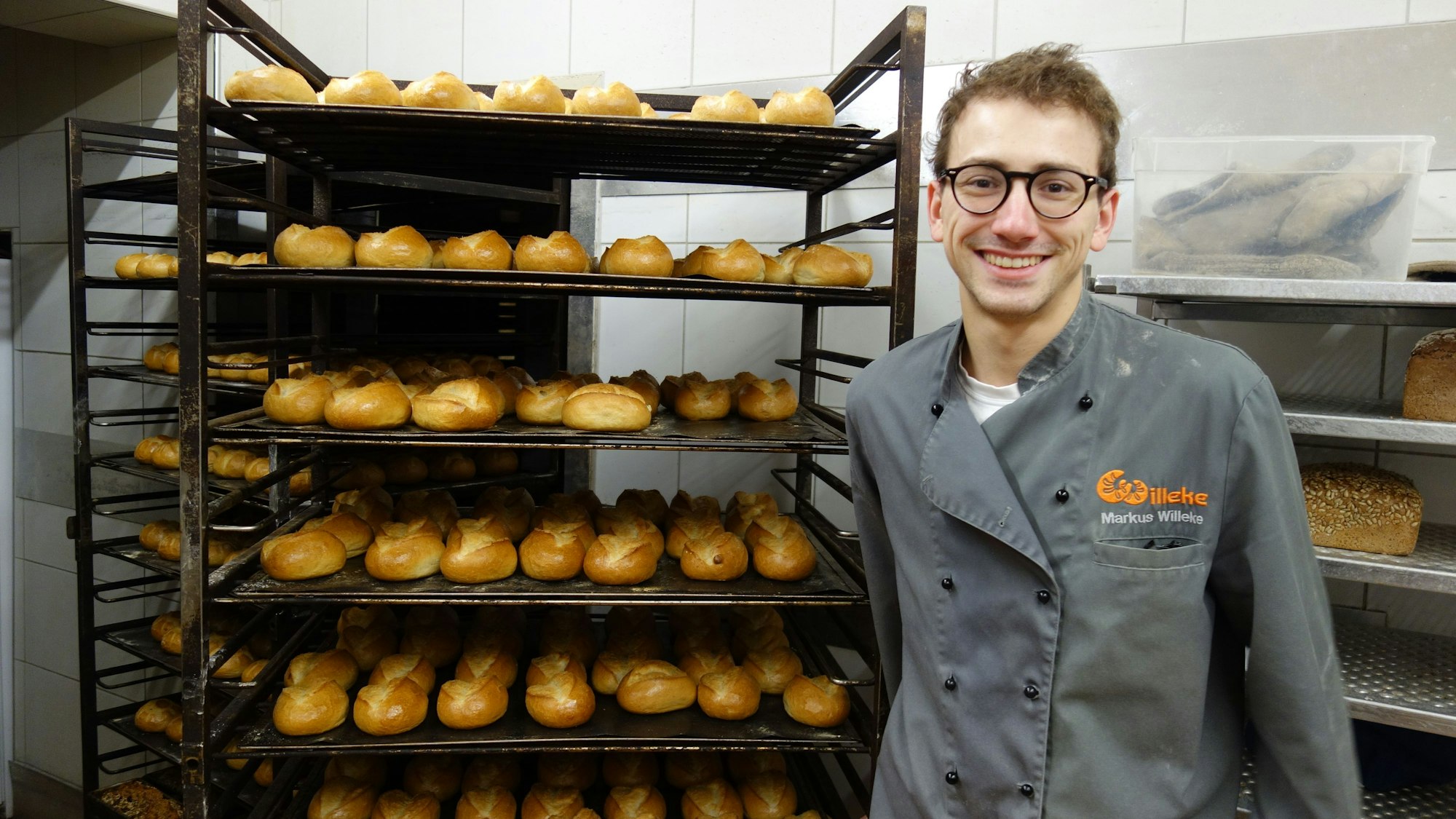 Ein Mitarbeiter der Bäckerei Willeke steht in der Backstube vor einem mit frischen Brötchen gefüllten Rostwagen.