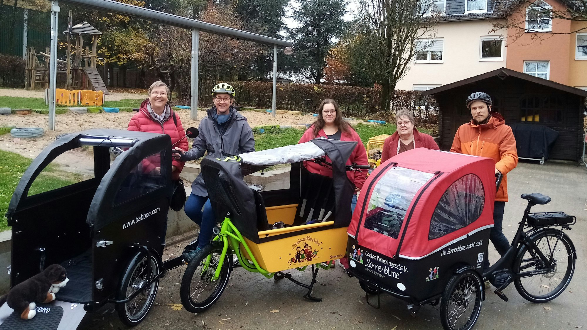 Vor einem Spielplatz befinden sich fünf Personen. Manche von ihnen sitzen auf Lastenrädern und tragen Fahrradhelme. Vor ihnen stehen drei Lastenräder in Schwarz, Gelb und Rot.