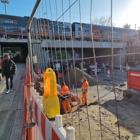 Blick auf die Baustelle an der Rathenaustraße am Bahnhof Leverkusen Mitte. Links gehen Fußgänger über Metallplanken, rechts daneben steht ein Bauzaun. Daneben stehen Bauarbeiter und rauchen. Ganz rechts fahren Autos auf der Rathenaustraße.