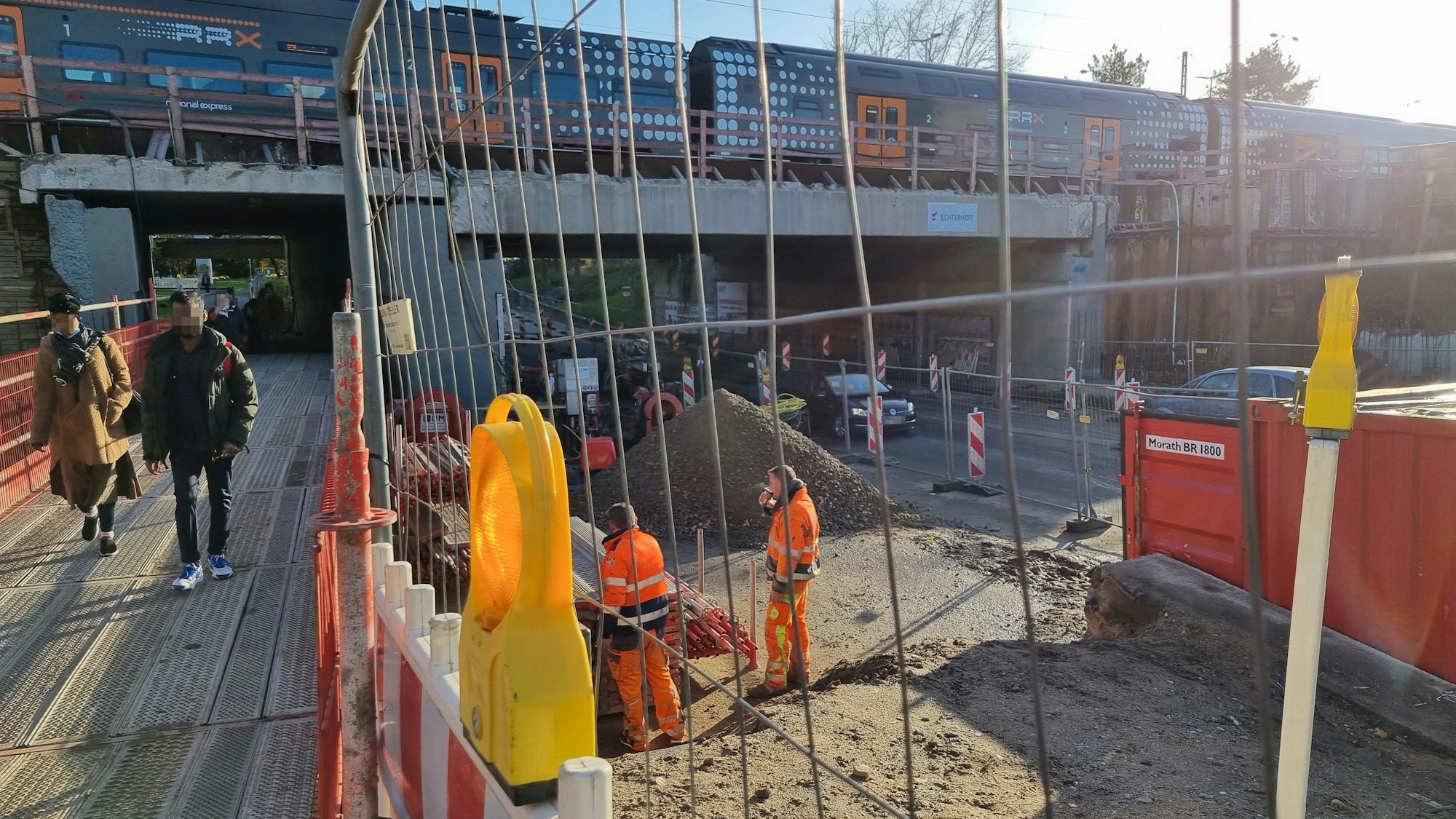 Blick auf die Baustelle an der Rathenaustraße am Bahnhof Leverkusen Mitte. Links gehen Fußgänger über Metallplanken, rechts daneben steht ein Bauzaun. Daneben stehen Bauarbeiter und rauchen. Ganz rechts fahren Autos auf der Rathenaustraße.