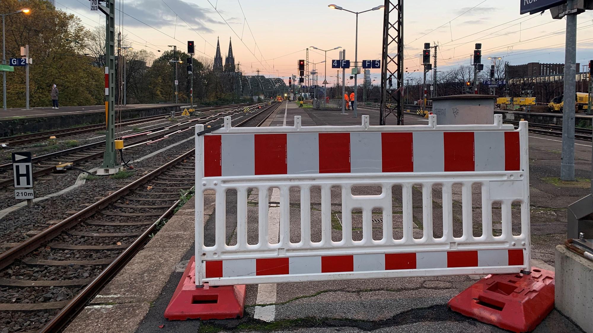 Eine Absperrung auf einem Bahnsteig des Bahnhofs Köln-Messe/Deutz ist zu sehen; dahinter in der Ferne die Spitzen des Kölner Doms.