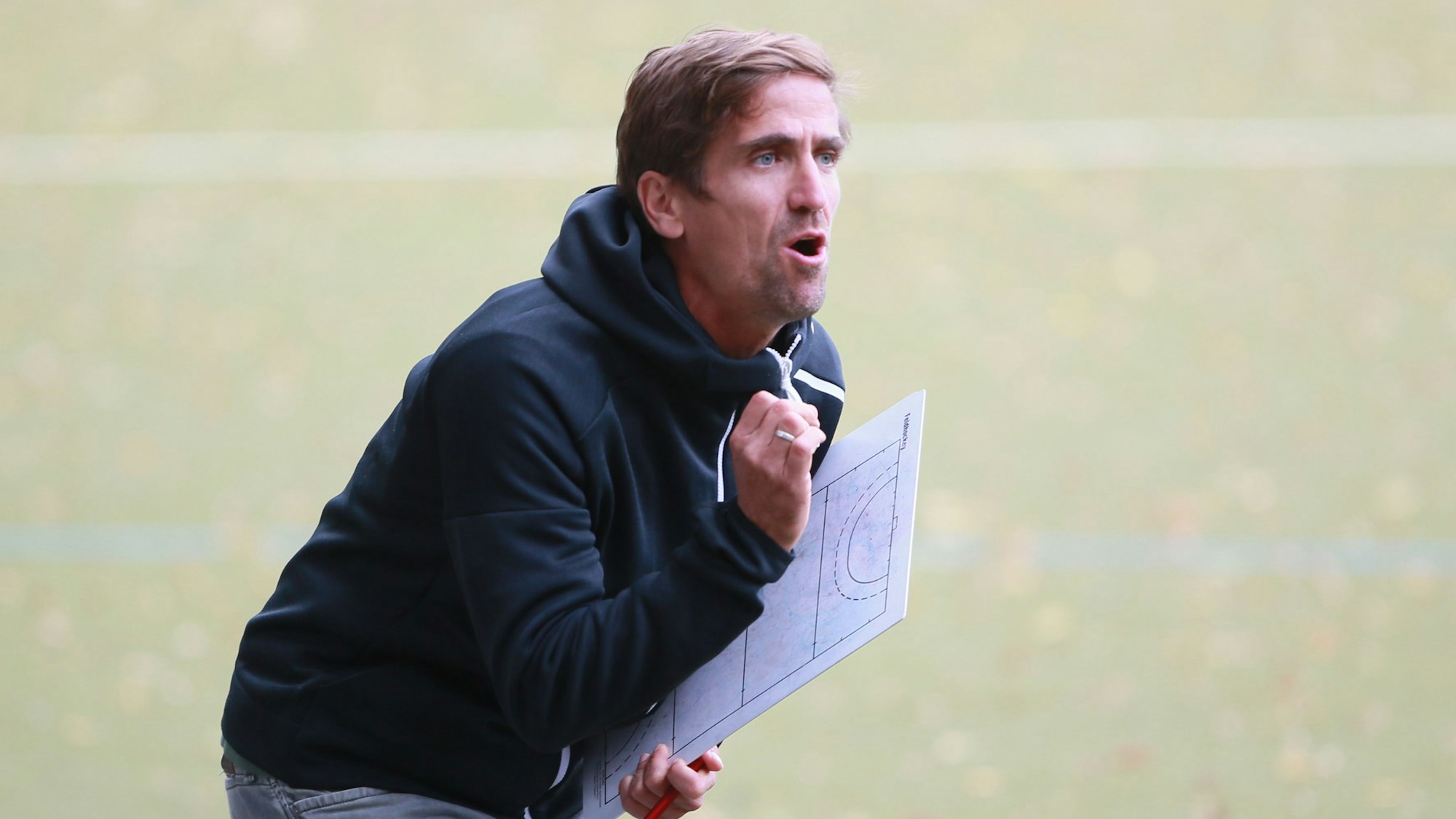 Ein Hockeytrainer steht am Spielfeldrand und gibt mit seinem Taktik-Brett in der Hand Anweisungen.