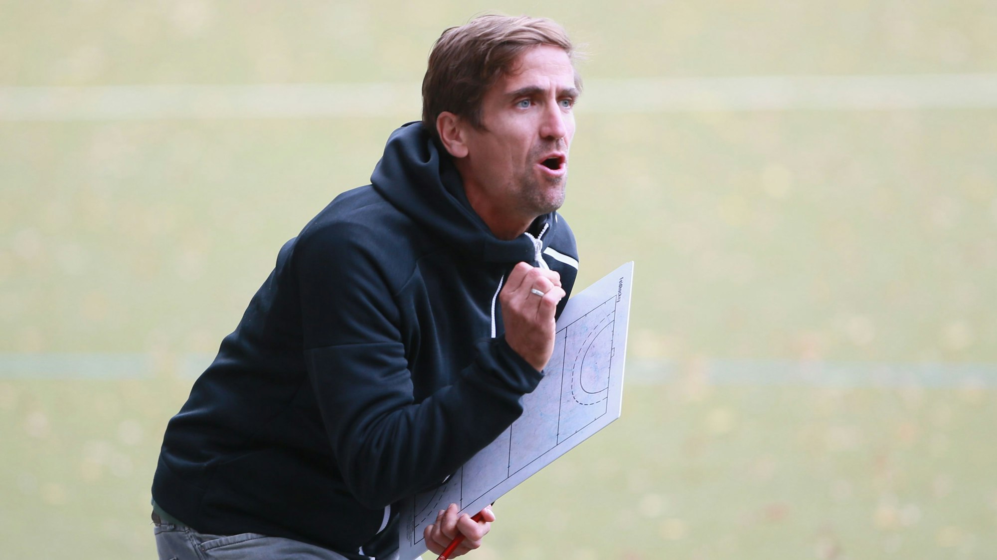 Ein Hockeytrainer steht mit seinem Taktikbrett am Spielfeldrand und gibt Anweisungen.