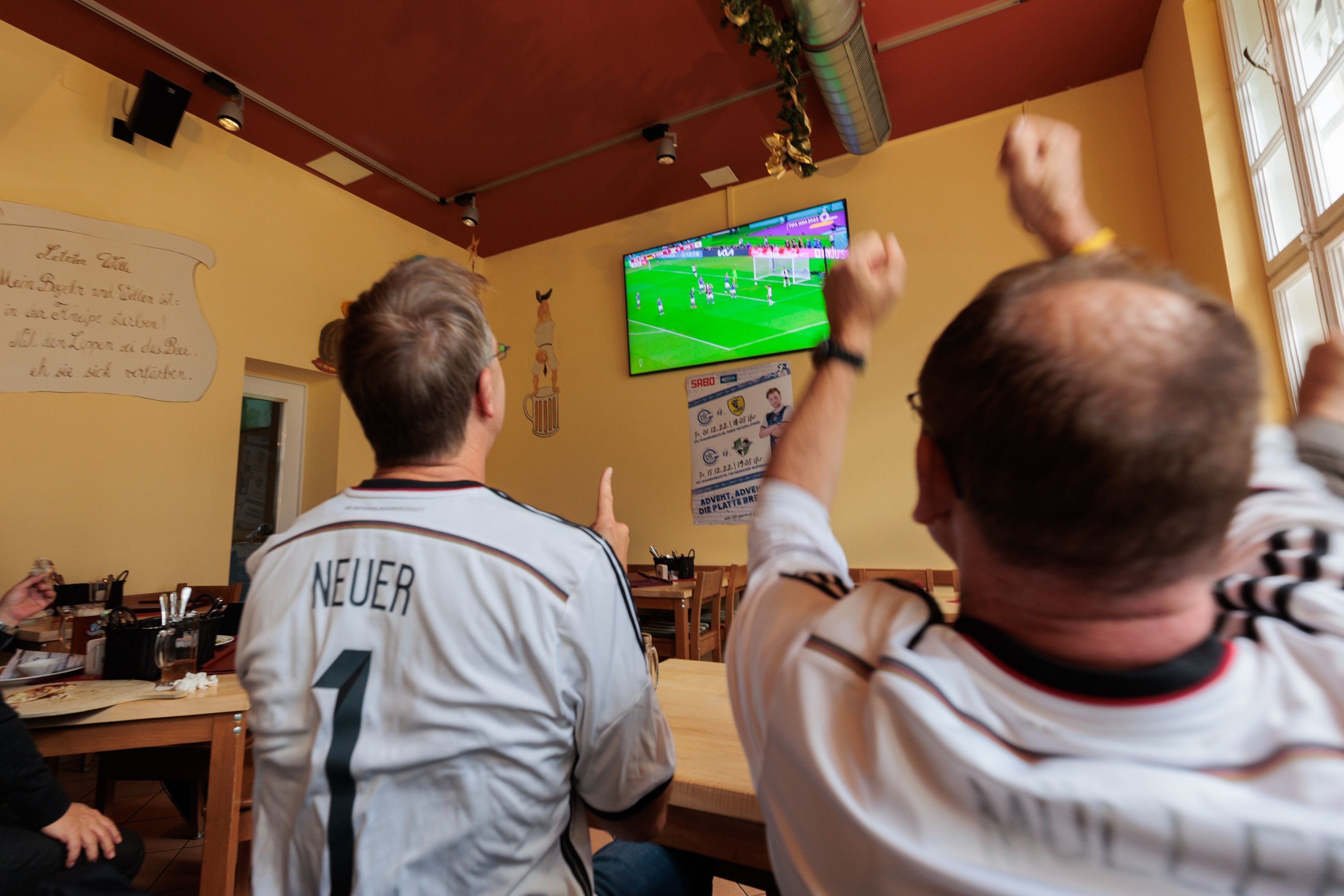 Zwei Fans stehen vor einem Bildschirm in einer Kneipe und freuen sich nach dem Tor für die deutsche Mannschaft.