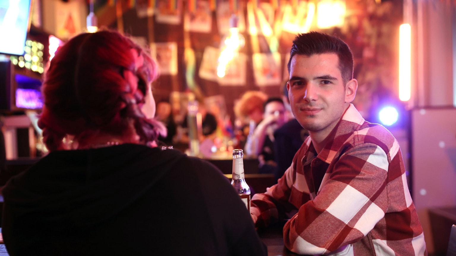 Zwei queere Jugendliche an der Bar des Kölner Jugendzentrums „Anyway“. Links mit dem Rücken zur Kamera sitzt Kitti, mit pinken Zöpfen. Daneben sitzt David im karierten Hemd und schaut in die Kamera.