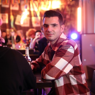 Zwei queere Jugendliche an der Bar des Kölner Jugendzentrums „Anyway“. Links mit dem Rücken zur Kamera sitzt Kitti, mit pinken Zöpfen. Daneben sitzt David im karierten Hemd und schaut in die Kamera.