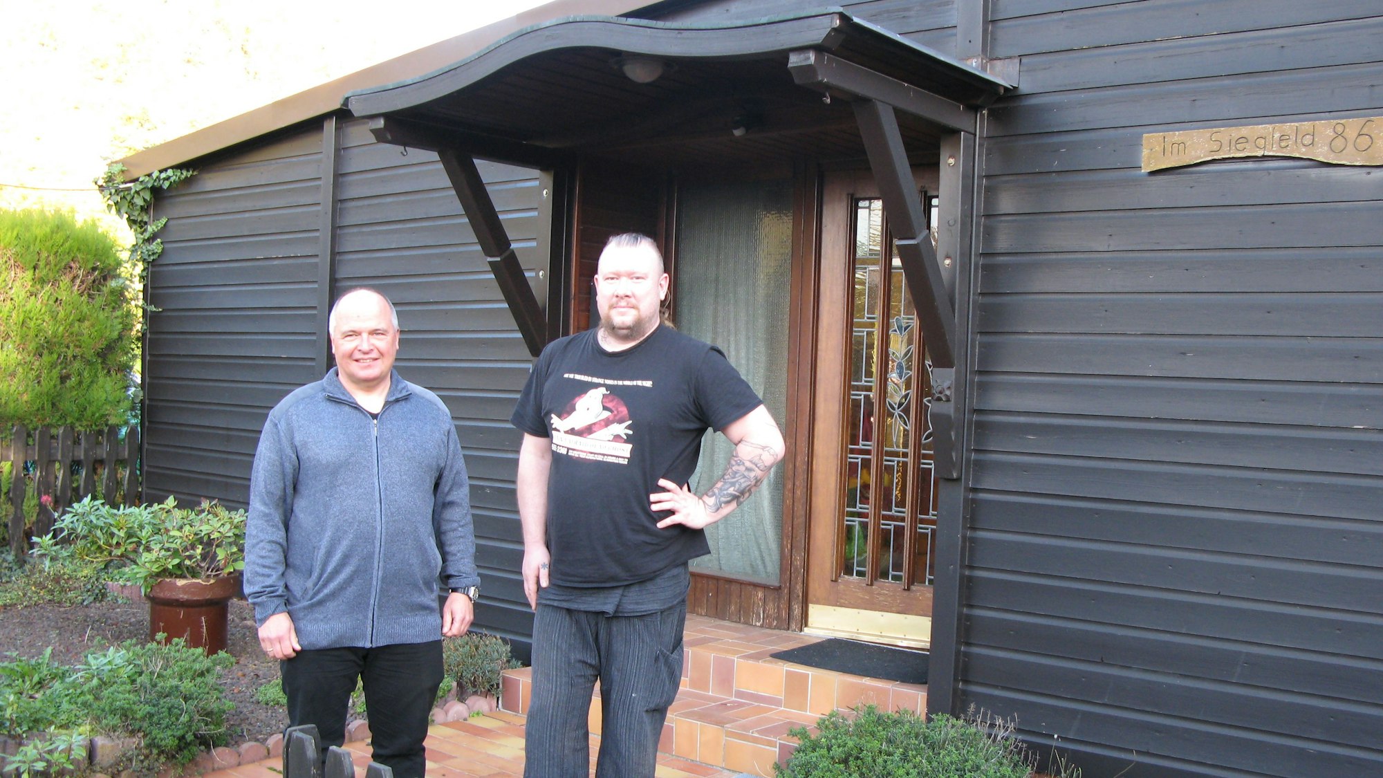 Vor dem mit Holz verkleideten Bungalow stehen zwei Männer und schauen in die Kamera.