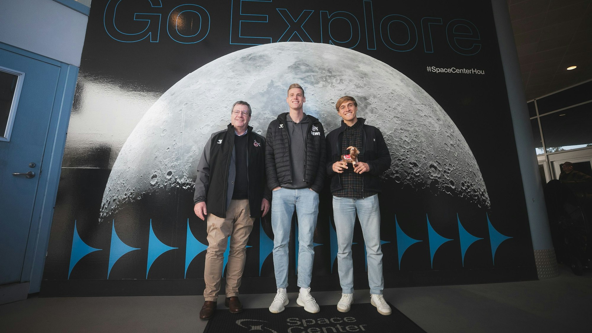 Werner Wolf, Steffen Tigges und Timo Hübers vom 1. FC Köln stehen vor einer großen Plakatwand im Space Center Houston der NASA. Darauf zu sehen ist er Mond mit der Aufschrift "Go Explore".