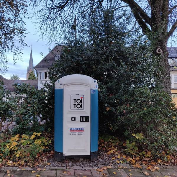 Eine blau-weiße Mobiltoilette steht unter Bäumen im Stadtpark am Busbahnhof Leichlingen