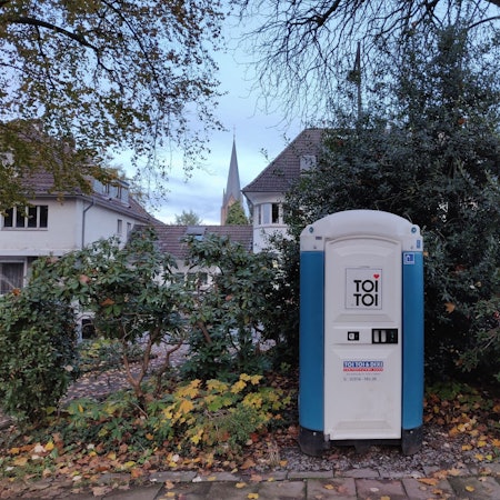 Eine blau-weiße Mobiltoilette steht unter Bäumen im Stadtpark am Busbahnhof Leichlingen