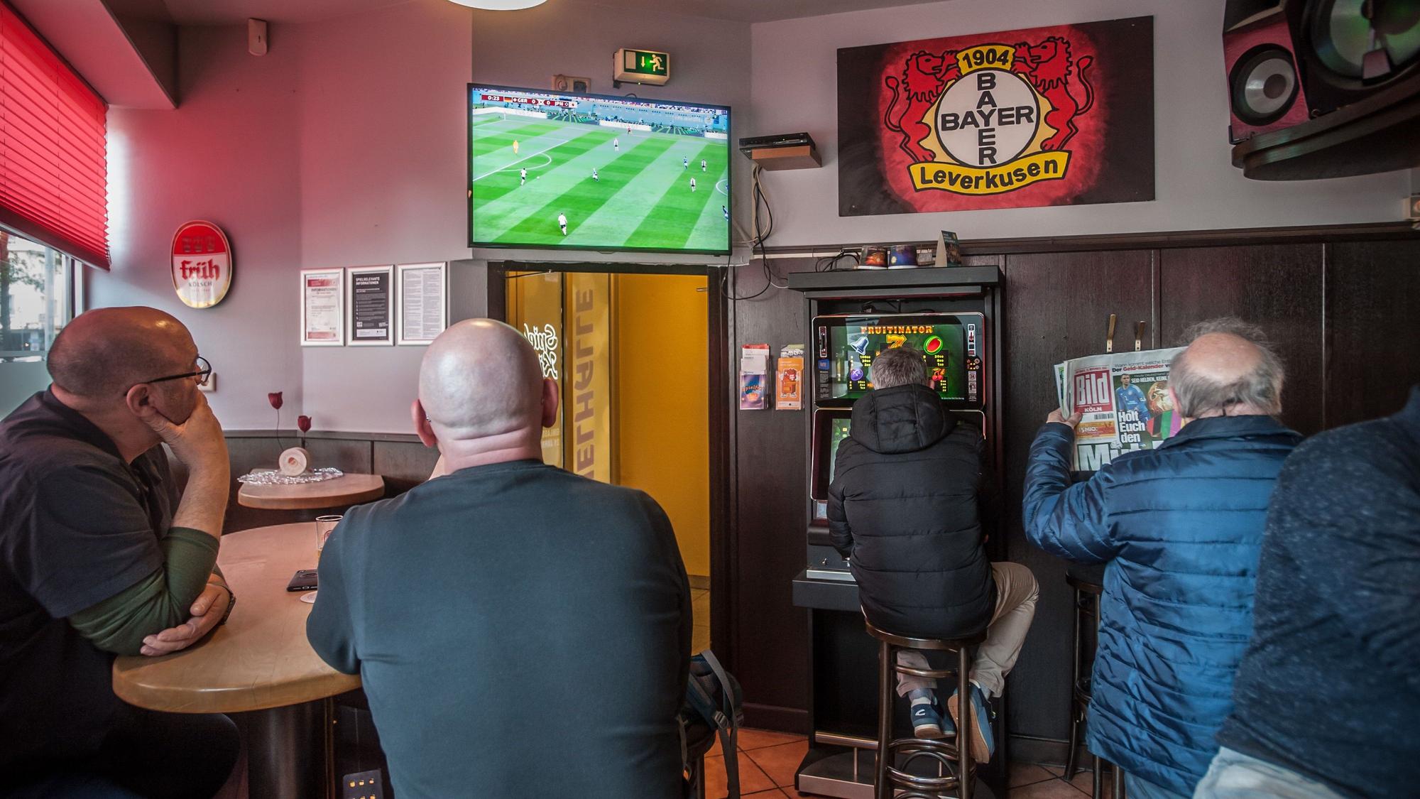 Blick auf vier Männer, die Fußball auf einem Fernseher schauen, einer sitzt am Spielautomaten.