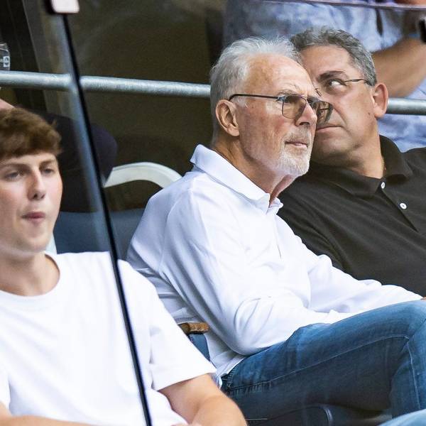 Franz Beckenbauer sitzt im Publikum bei einem Fußballspiel. Neben ihm ein Mann. Beckenbauer trägt eine getönte Brille und schaut mit ernster Miene auf das Spielfeld. Er trägt ein weißes Oberteil.