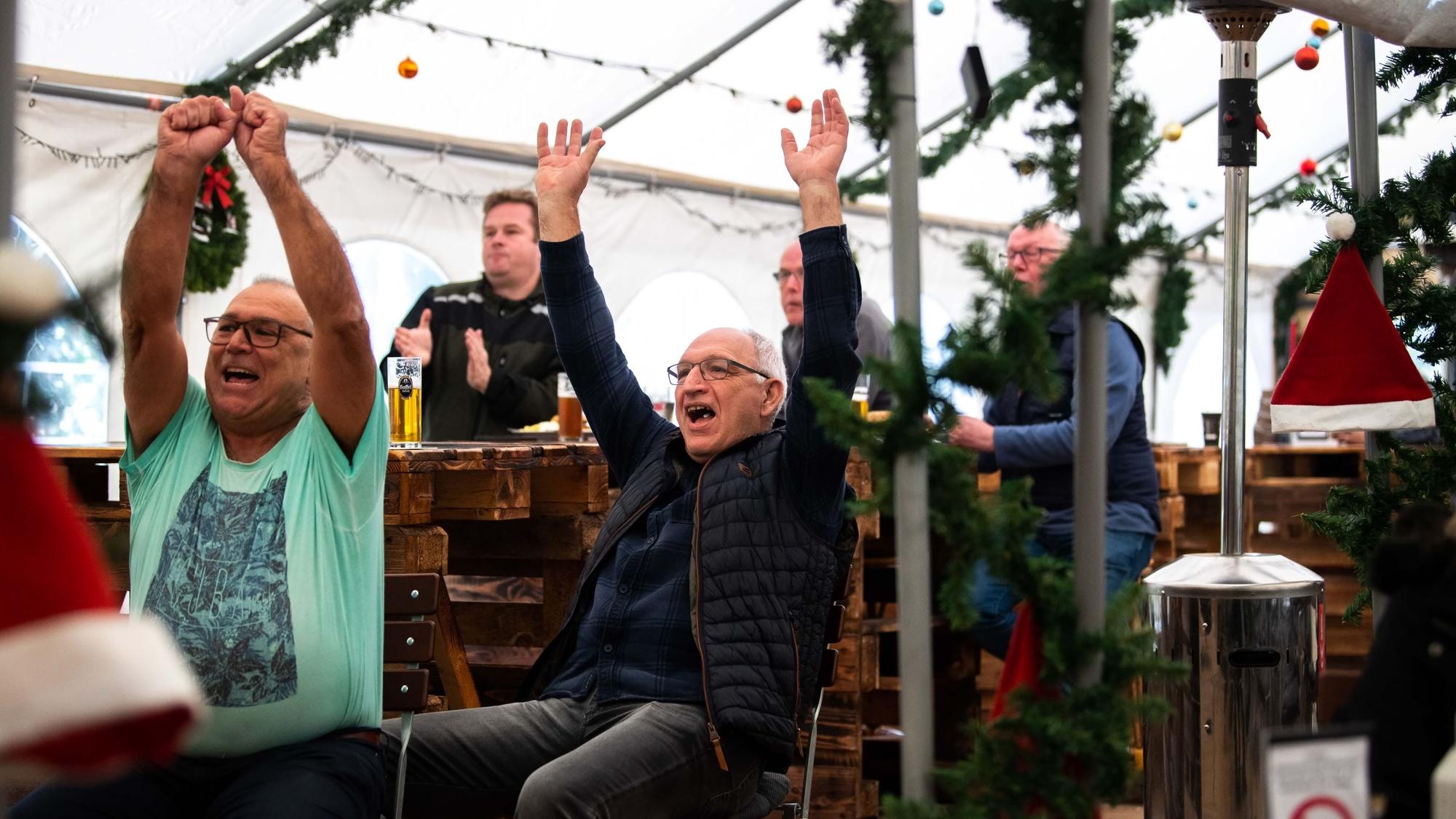 Fußballfans im Biergarten in Kommern werfen jubelnd die Arme in die Höhe und freuen sich über das 1:0 der deutschen Nationalmannschaft gegen Japan. Im Zelt im Biergarten hängt Weihnachtsdekoration.